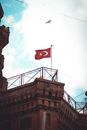 From below of aged brick house with metal fence on roof and wavy flag representing crescent and star under sky with clouds and flying bird in city