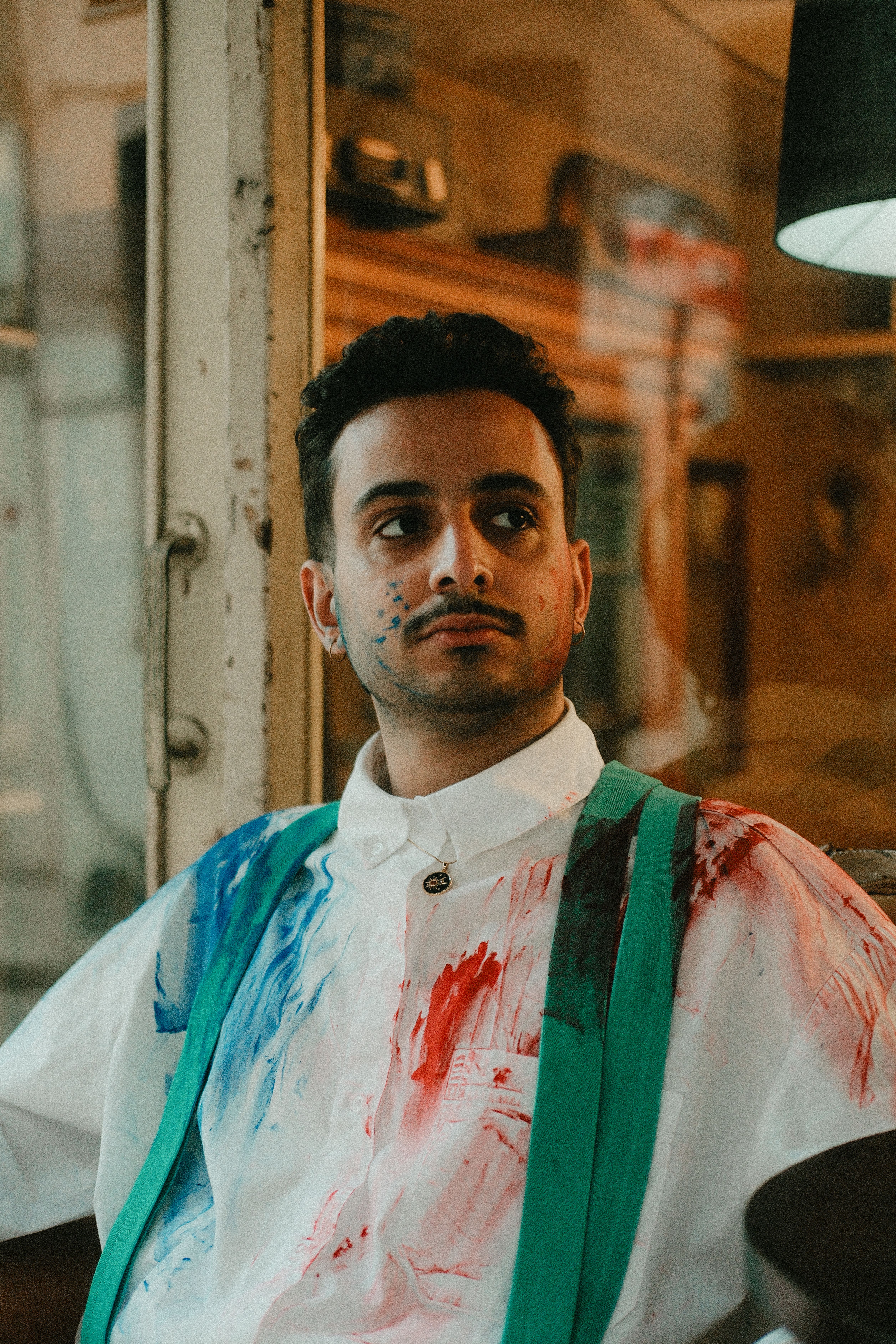 Young artist with paint-splattered shirt sits thoughtfully in an indoor setting.