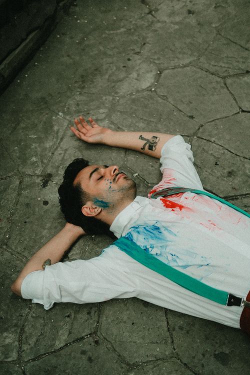Free A young man lies on a paved surface with colorful paint splashes on his shirt. Stock Photo