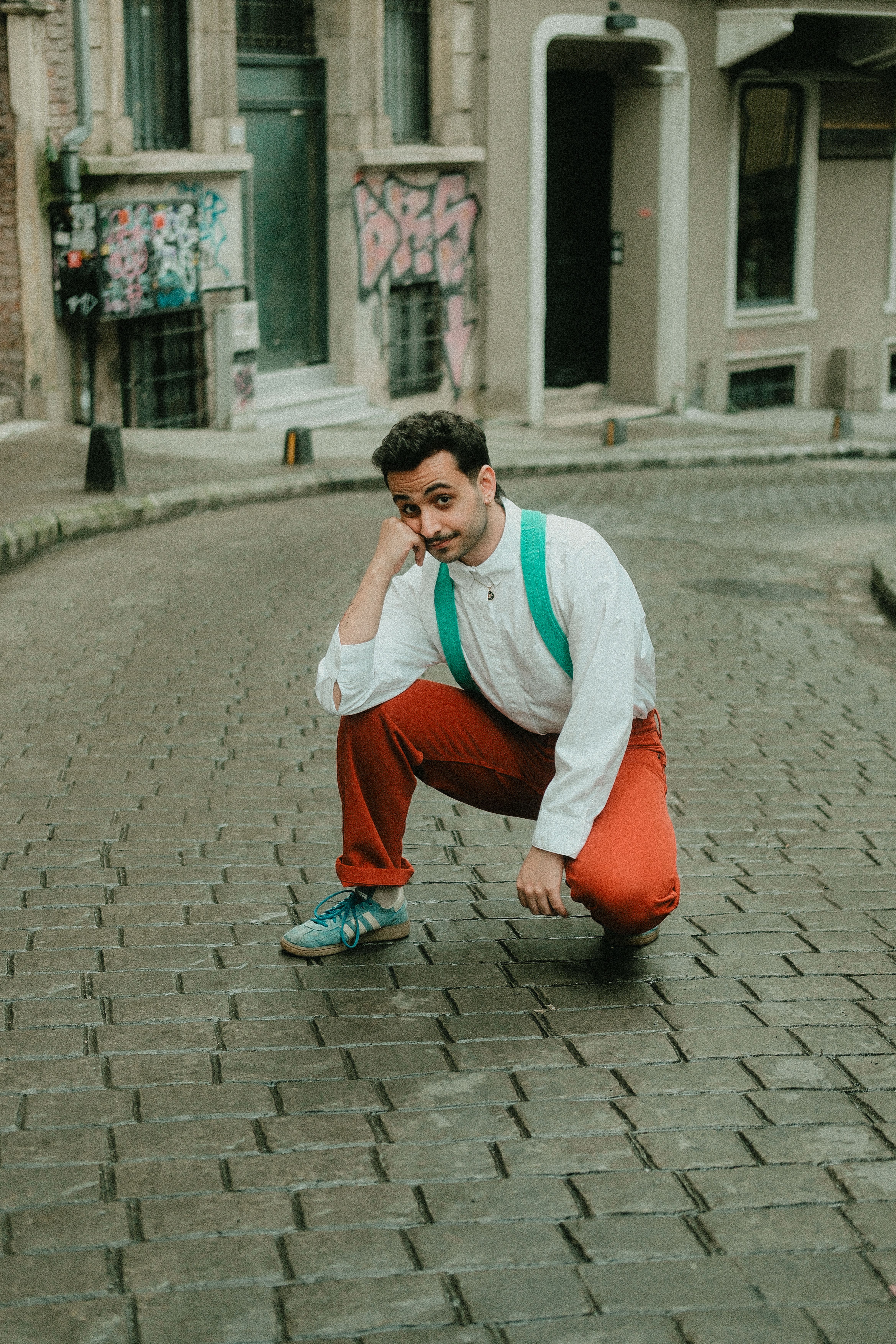 A stylish young man kneels on a cobblestone street, surrounded by urban graffiti.