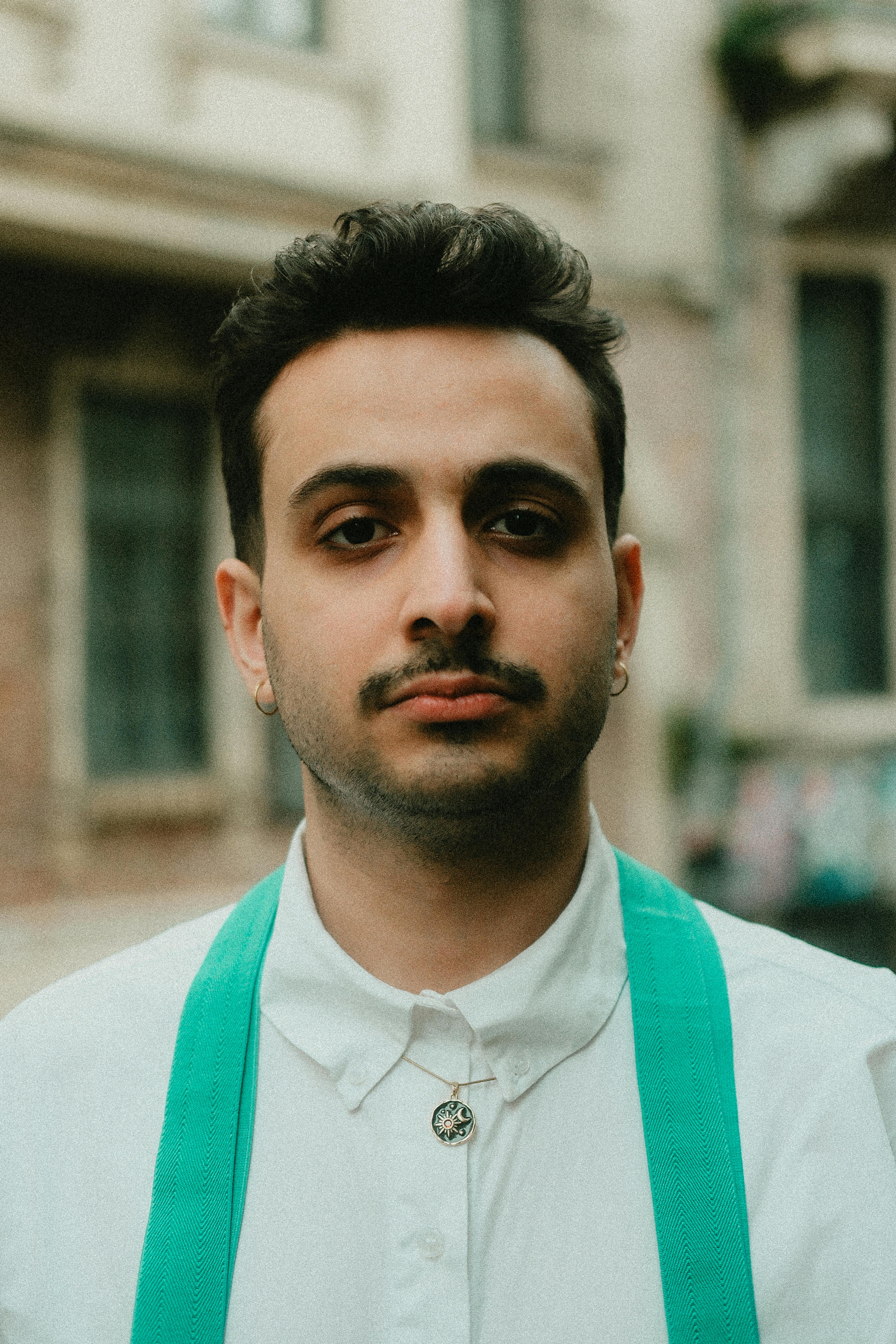 Close-up portrait of a young man wearing green suspenders, standing outdoors with a neutral expression.
