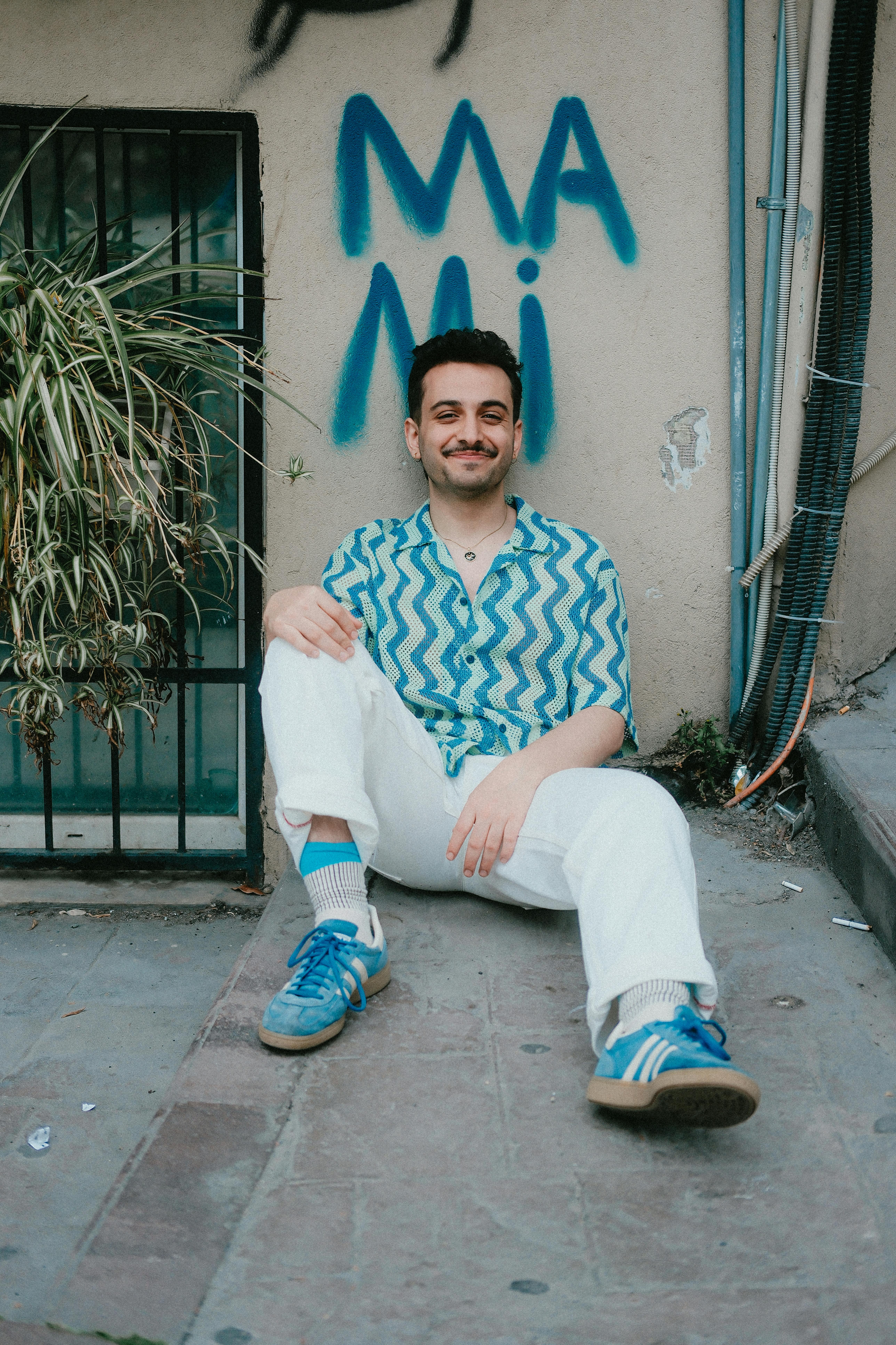 Man in casual attire sitting on a pavement near graffiti, expressing relaxed mood.