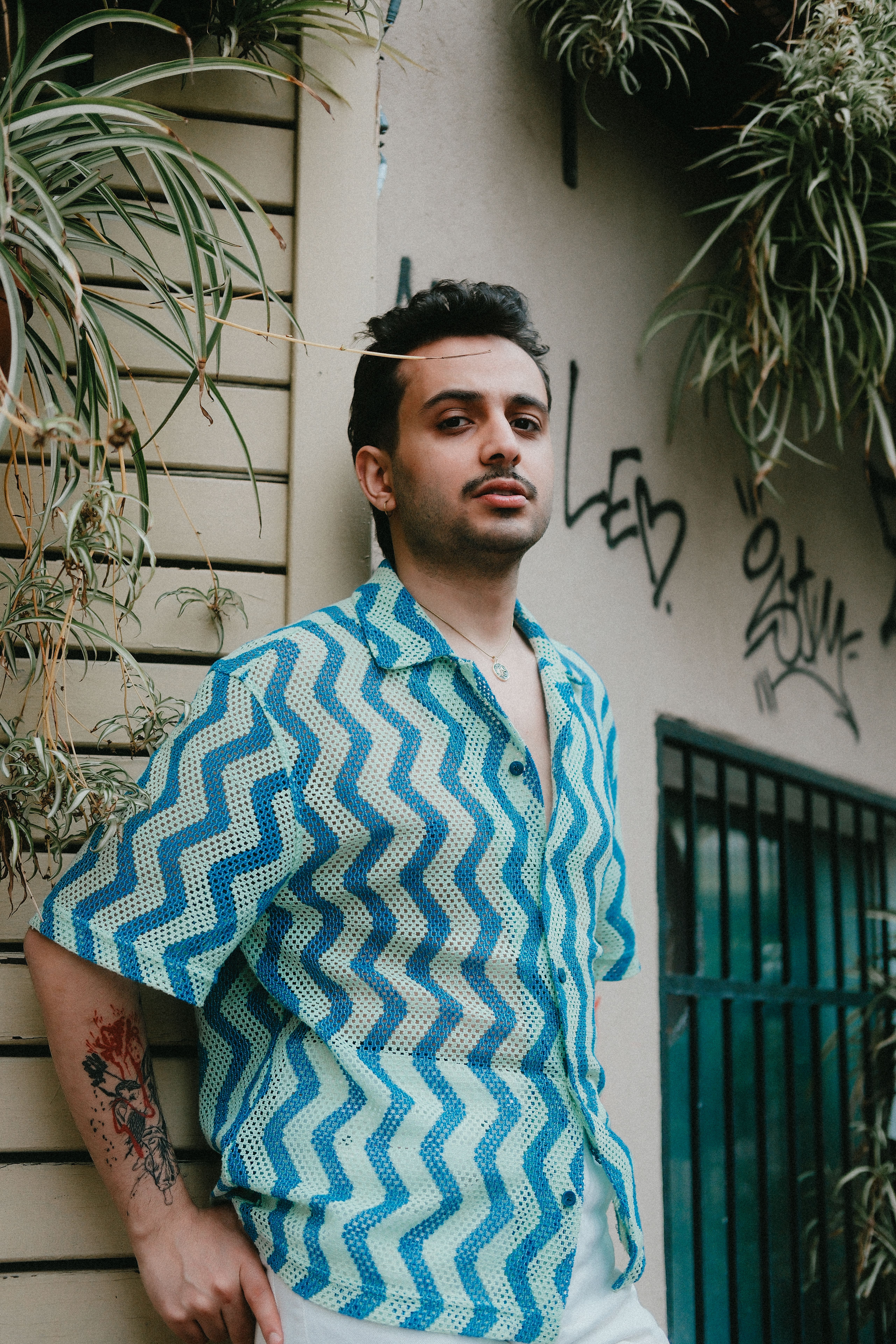Stylish young man in patterned shirt posing by a graffiti-covered wall with plants.