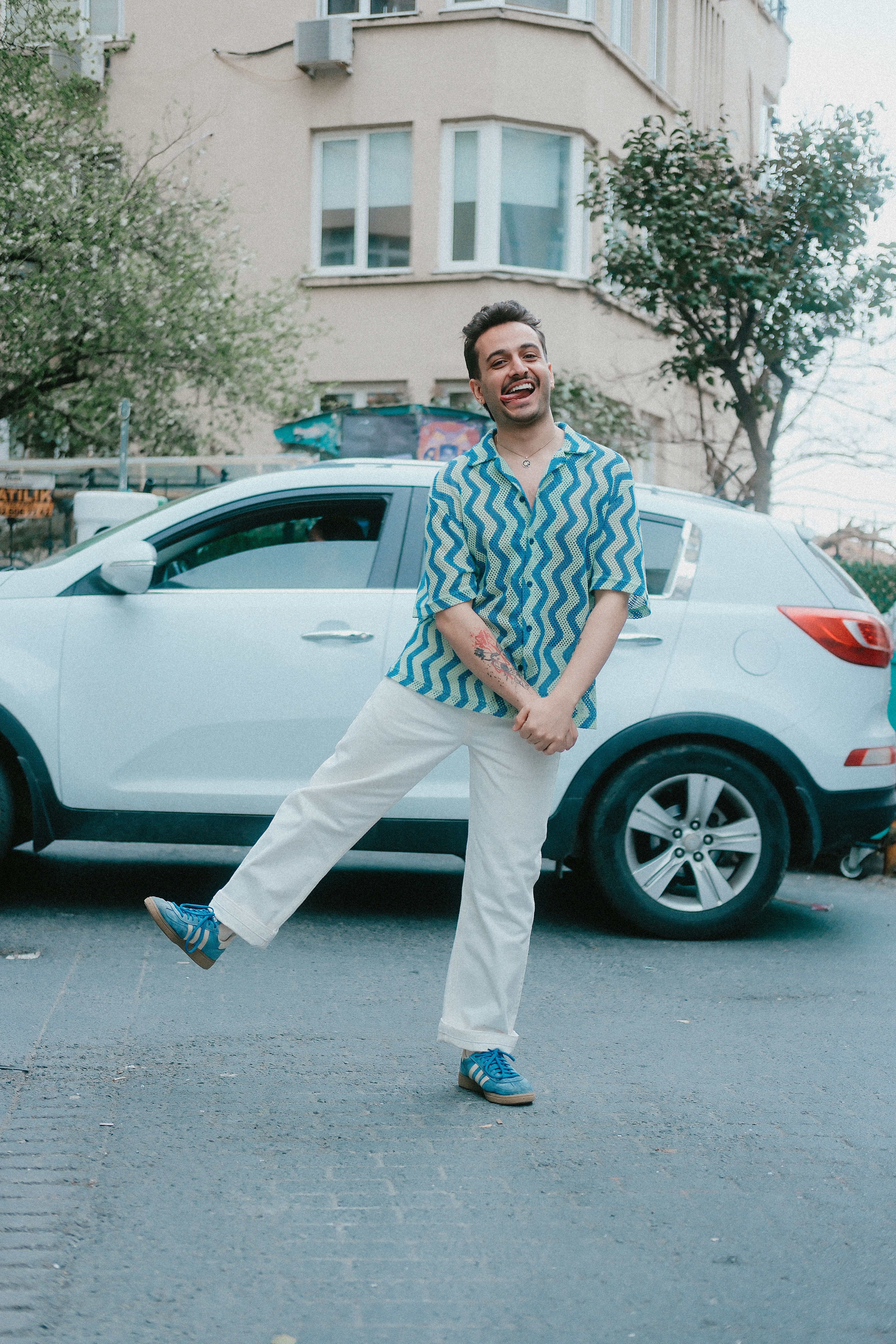 Cheerful man in blue patterned shirt playfully poses near a white car on a city street.