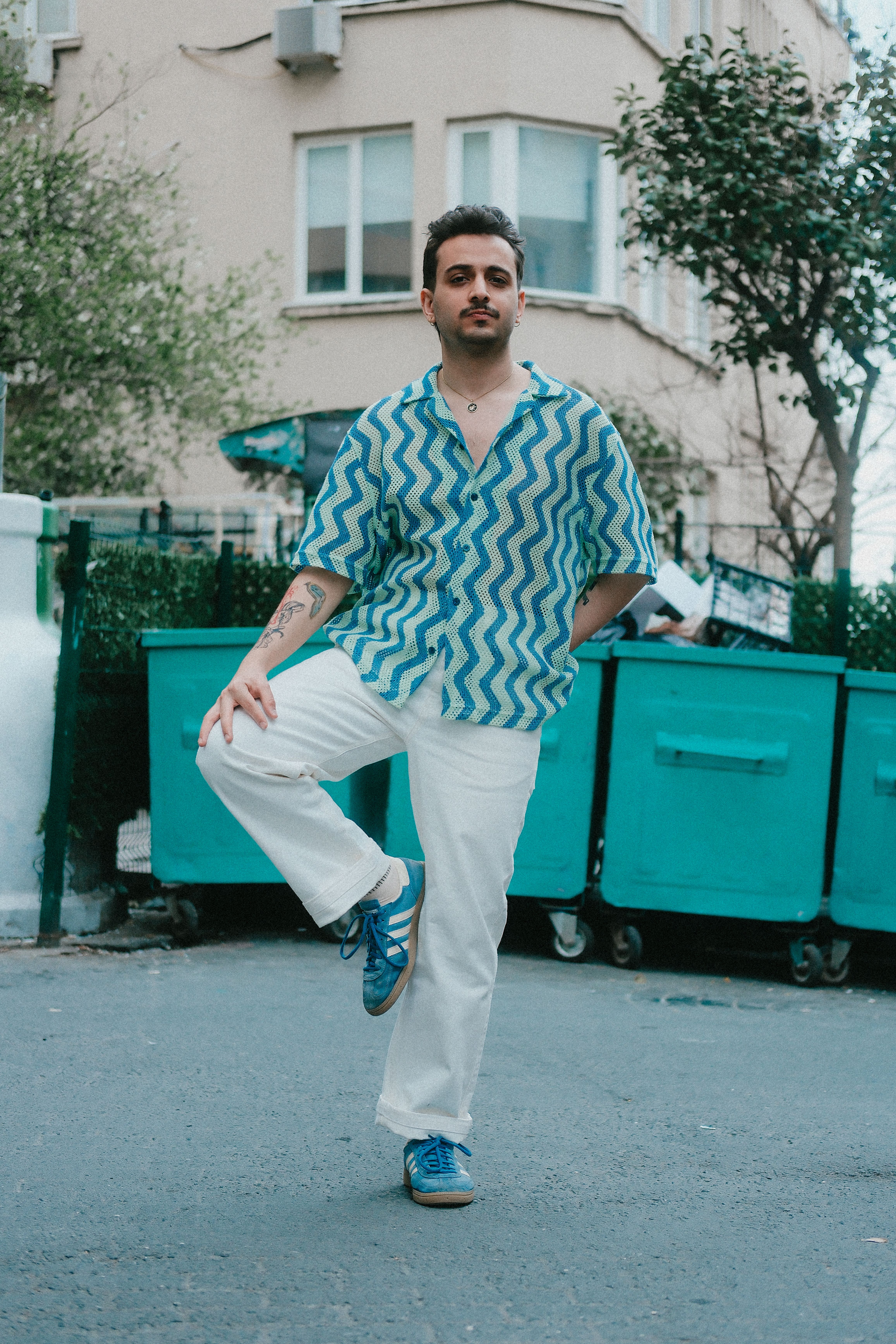 Fashionable man poses stylishly on a city street with dumpsters in the background.