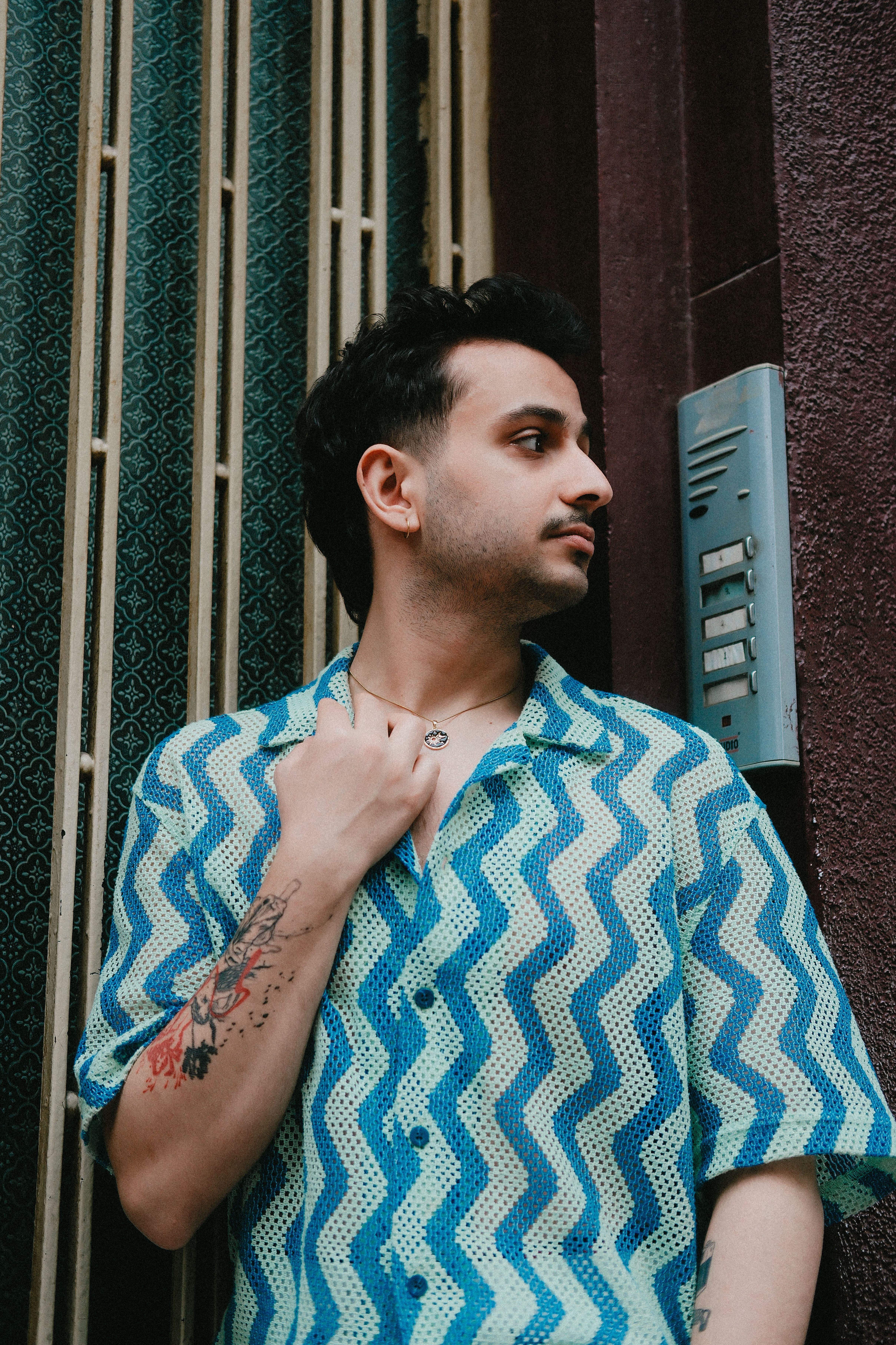 Young man with tattoos posing outdoors near an intercom system.