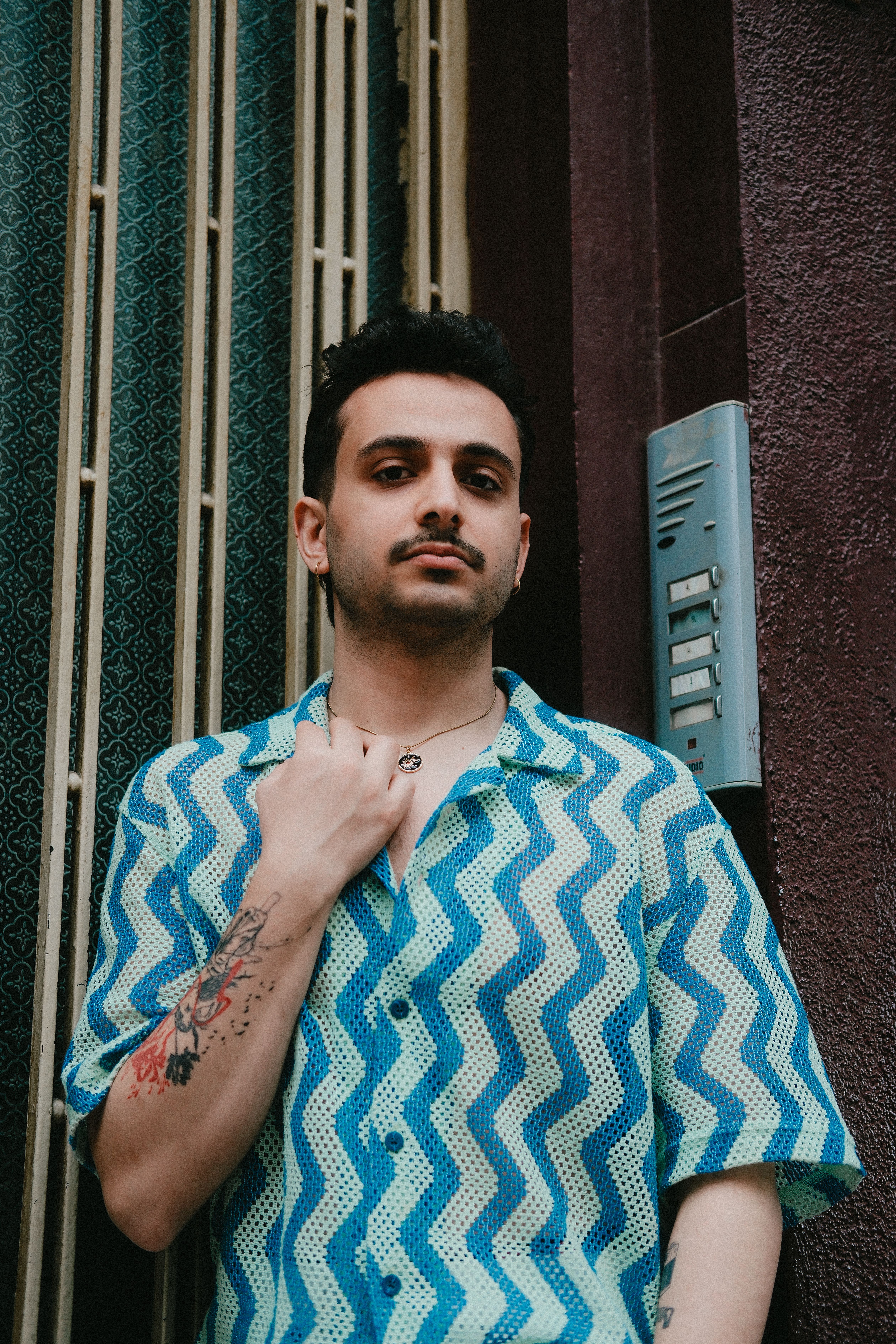 A young adult with tattoos poses confidently in a vibrant patterned shirt outdoors.