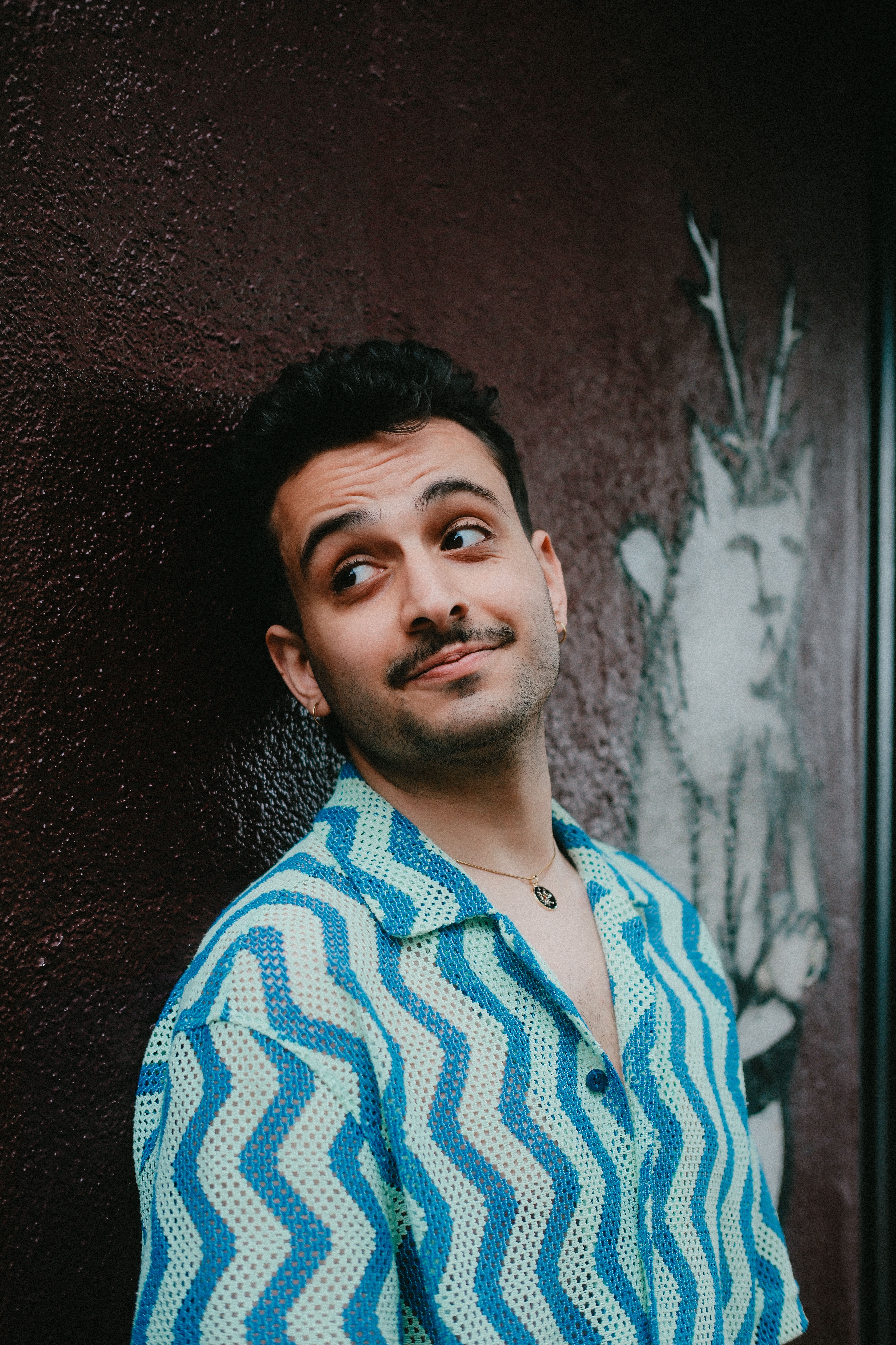 A man with a mustache smiles playfully against a textured, artistic wall.