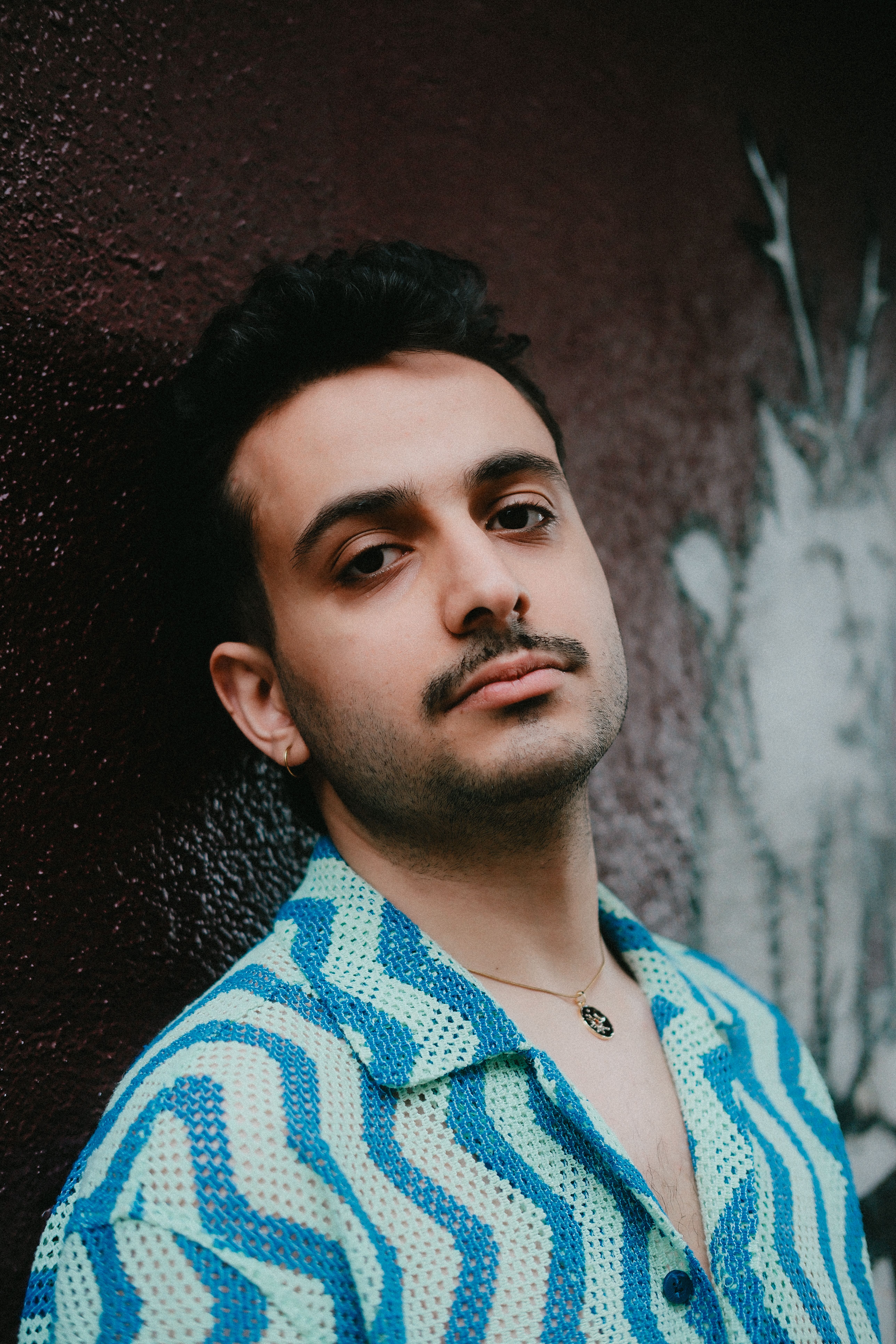 Young man with mustache in a blue patterned shirt leaning against a dark wall.