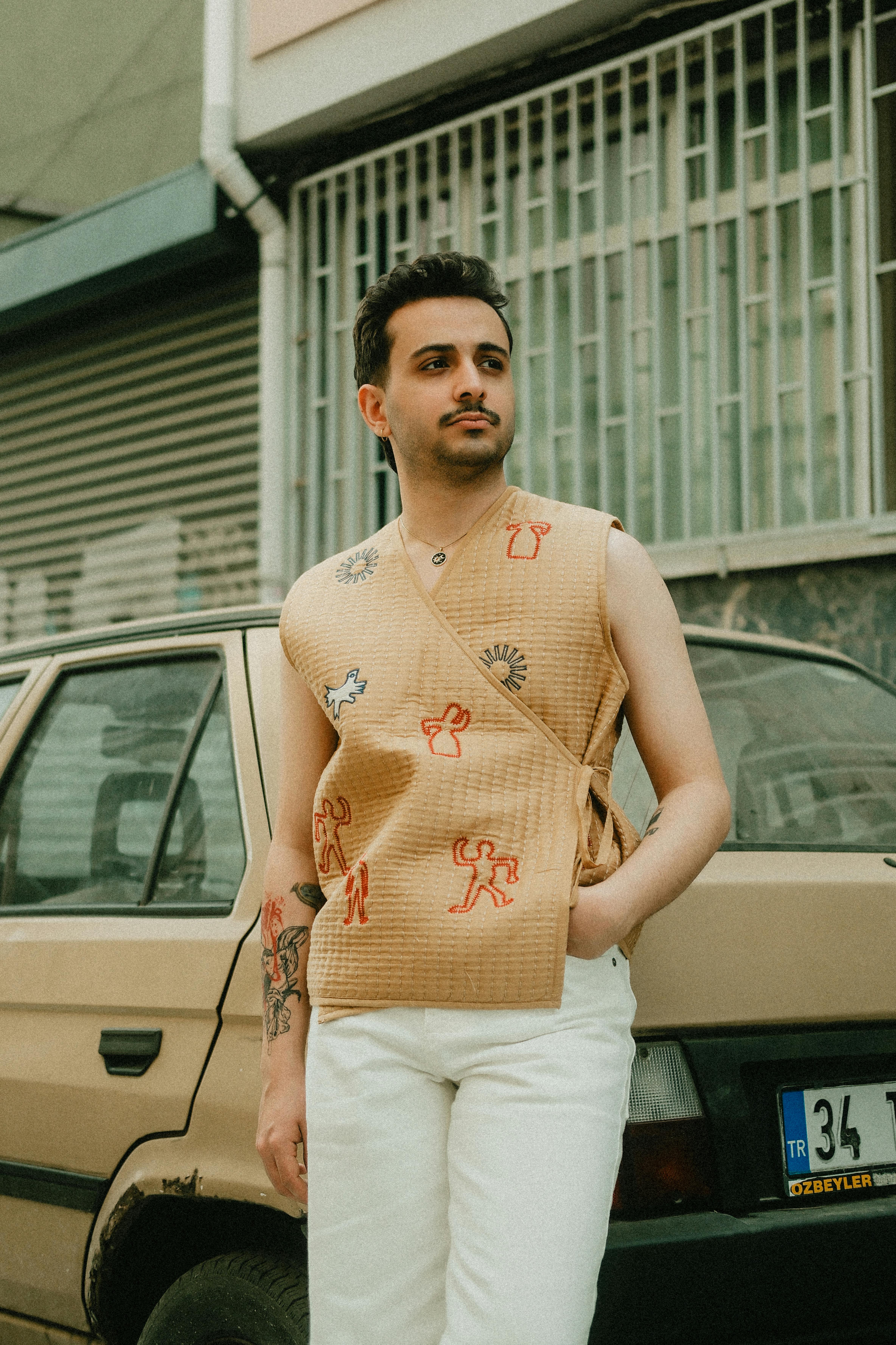 A fashionable young man in a patterned vest stands by a retro car.