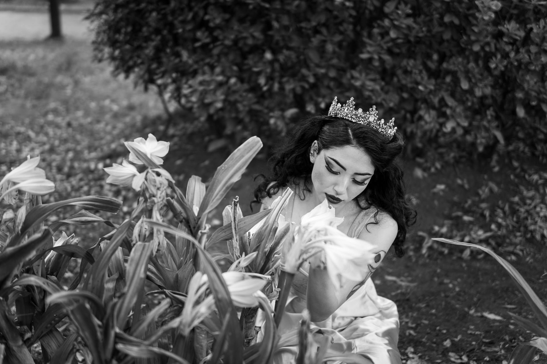 Black and white portrait of a woman in a princess costume with a tiara, sitting among flowers in a garden.