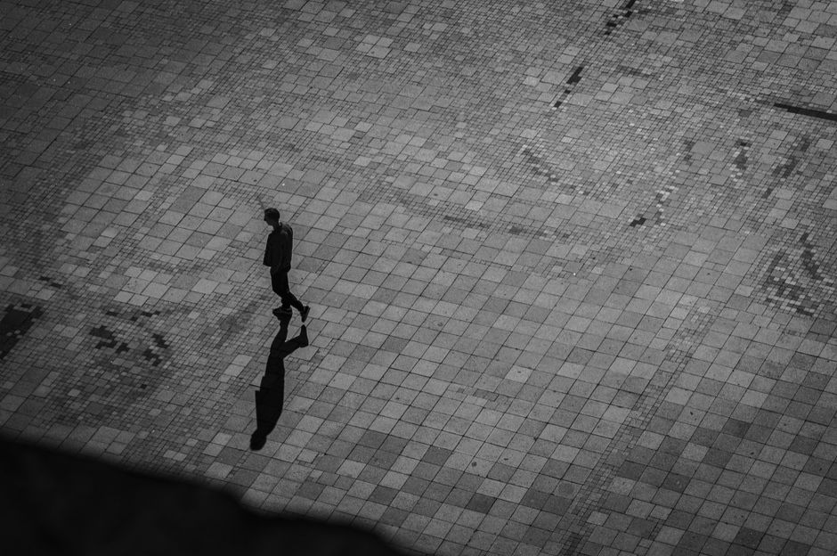 A solitary person walks on a patterned tiled square, casting a distinct shadow.