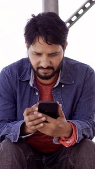 Casually dressed man focused on smartphone while seated outdoors, suggesting modern technology use in everyday life.
