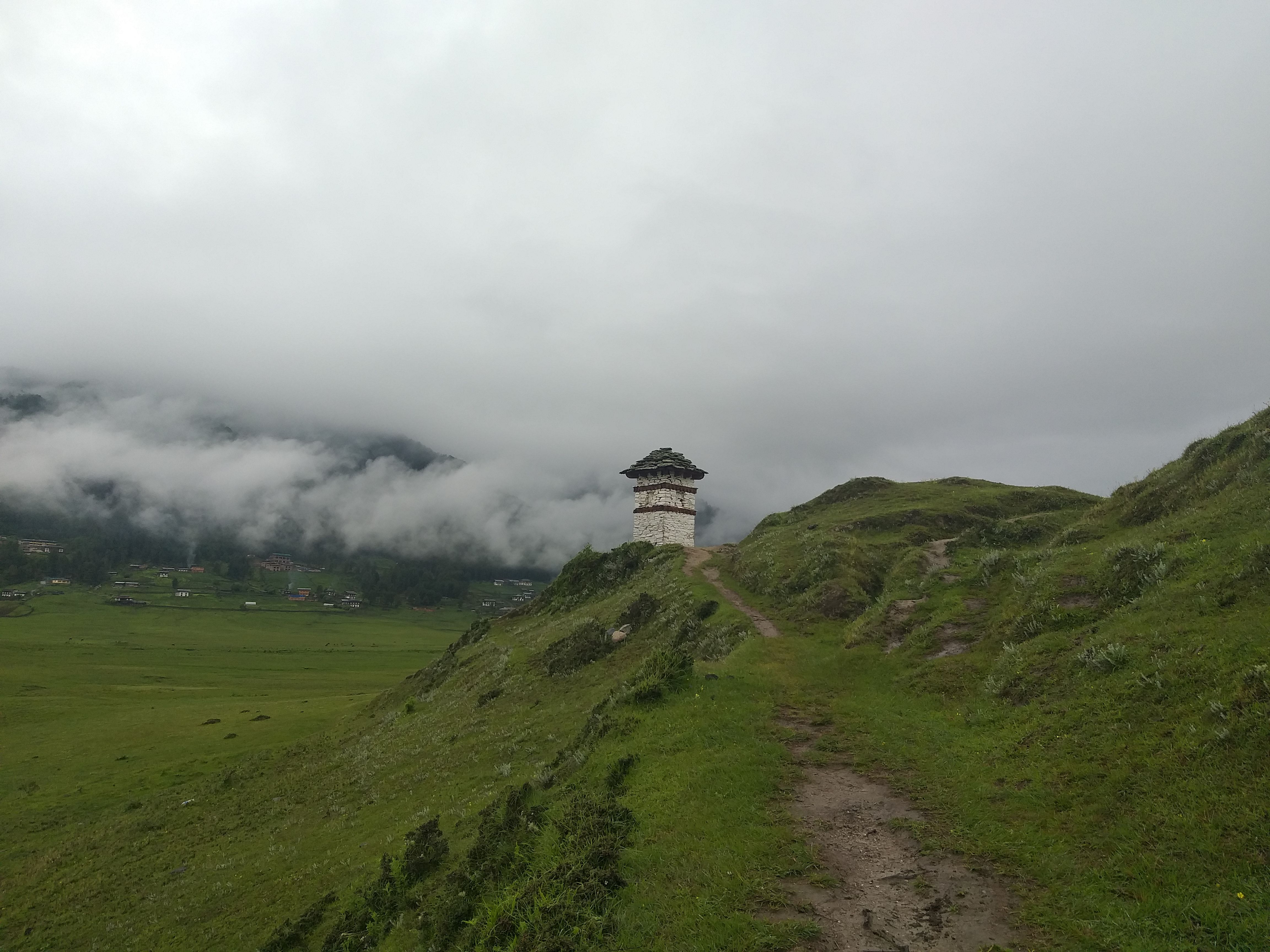 Un sendero que sube por una loma de hierba hasta una pequeña torre blanca sobre un valle amplio con niebla, en Wangdue Phodrang, Bután