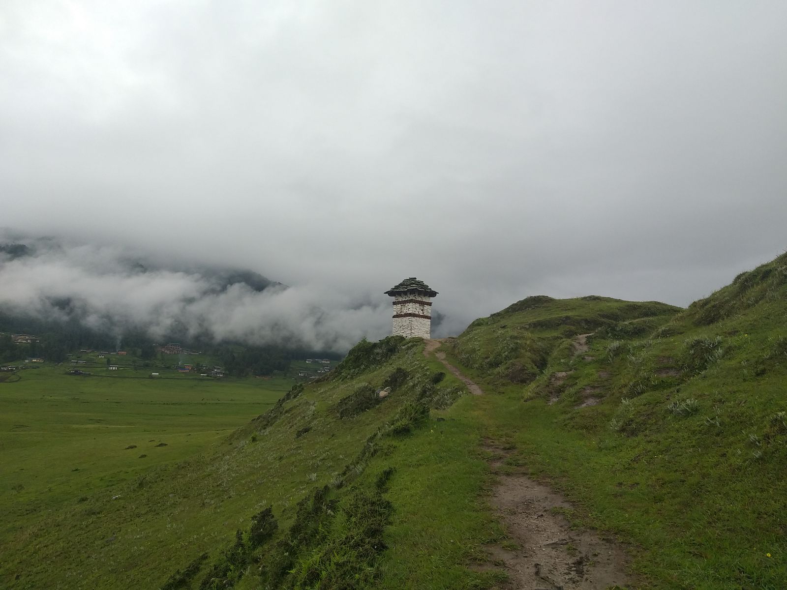 A footpath climbing a grassy ridge to a small white tower above a wide misty valley floor in Wangdue Phodrang, Bhutan