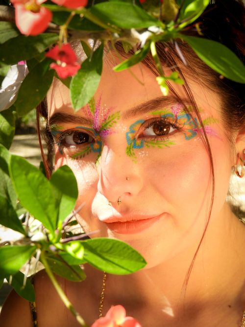 Free A woman with intricate floral face paint surrounded by green leaves, capturing a vibrant spring essence. Stock Photo