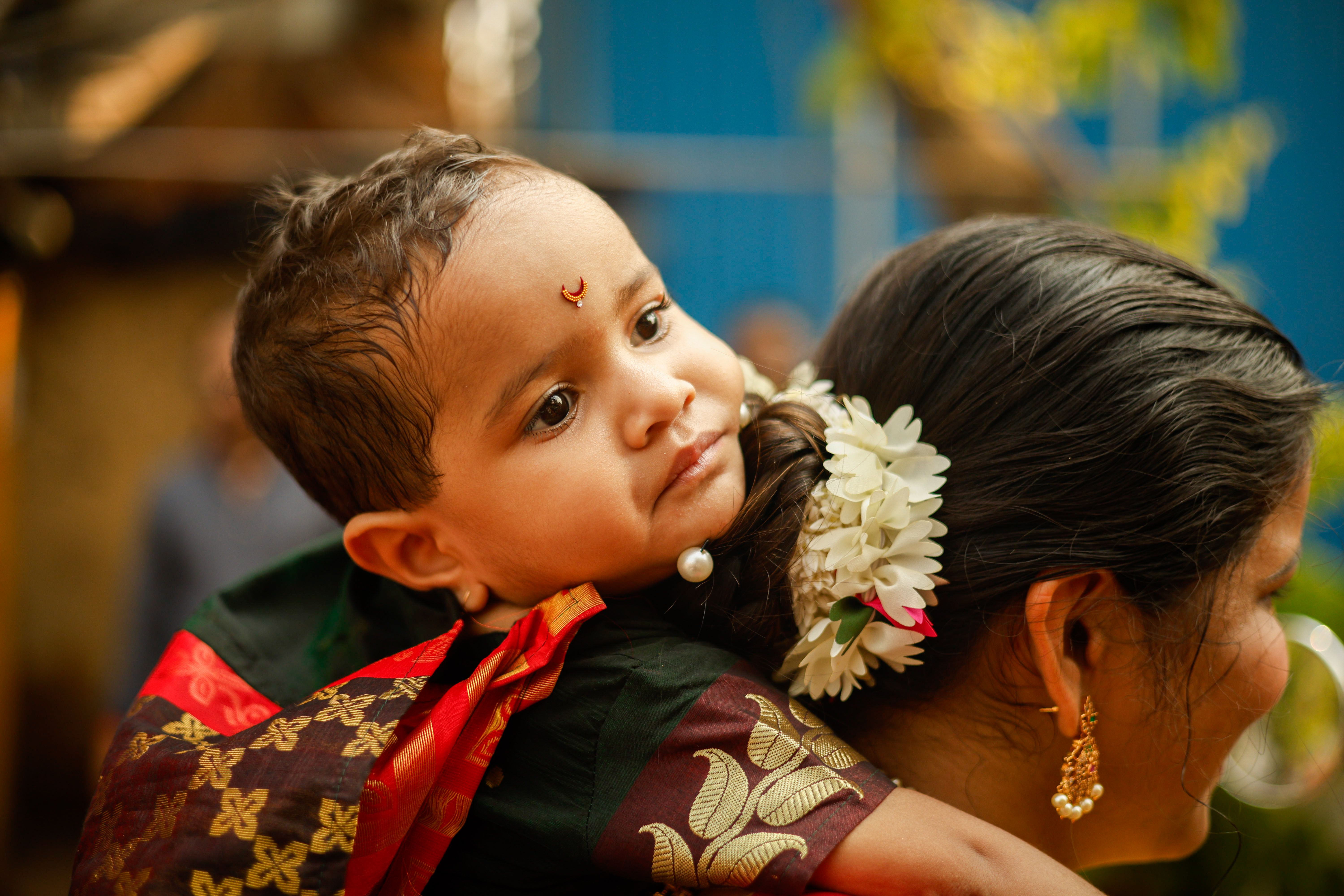 A close-up of a baby in traditional clothing resting on a woman's shoulder with floral hair adornment.