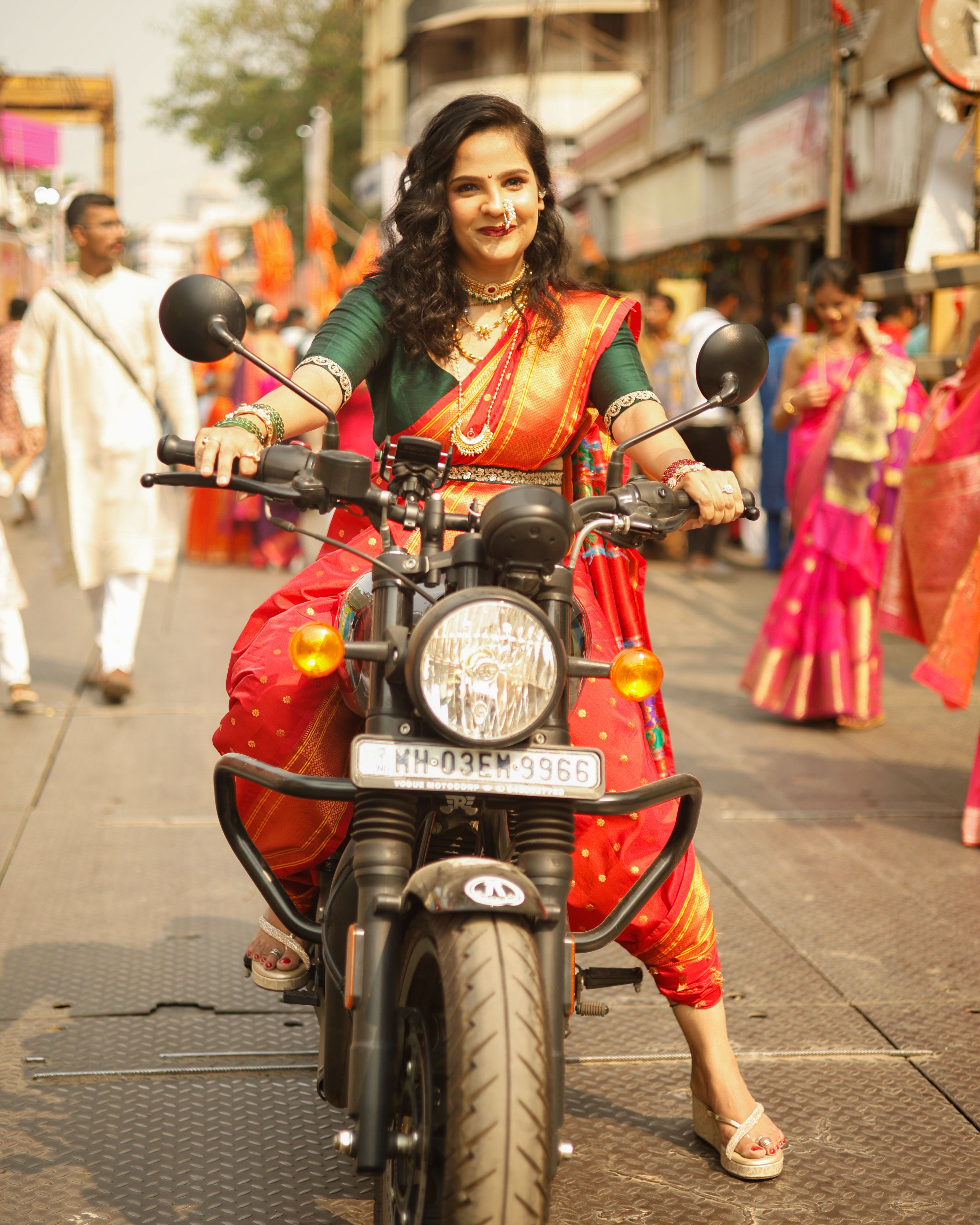 Woman in a vibrant saree rides a motorcycle during a lively festival with colorful street surroundings.