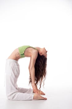 A woman in a green top and white pants performs yoga camel pose on a white background.