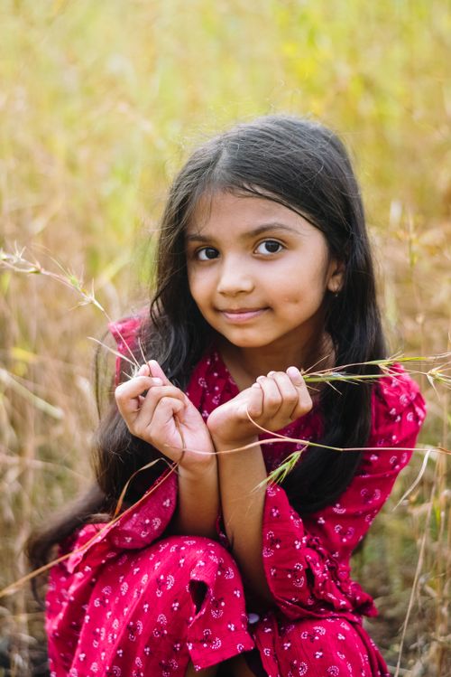 Free Portrait of a smiling young girl in a vibrant red dress amidst tall grass in Navi Mumbai. Stock Photo