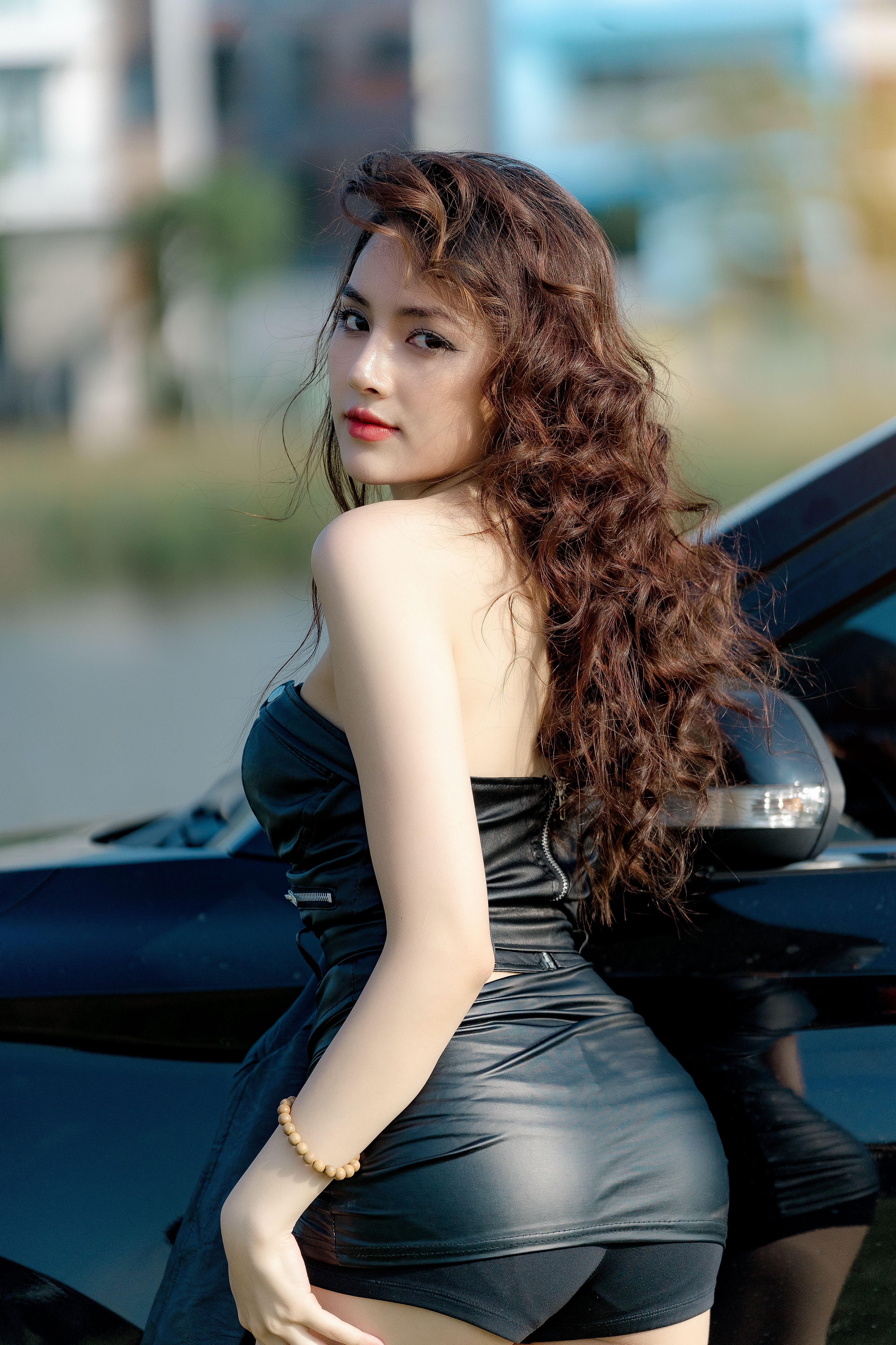 Confident woman in black attire poses beside a sleek car on a sunny day.