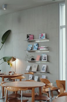 A stylish modern cafe interior featuring a wall bookshelf, wooden tables, and a potted plant.