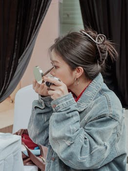 Young woman in denim jacket applying makeup indoors using a compact mirror.