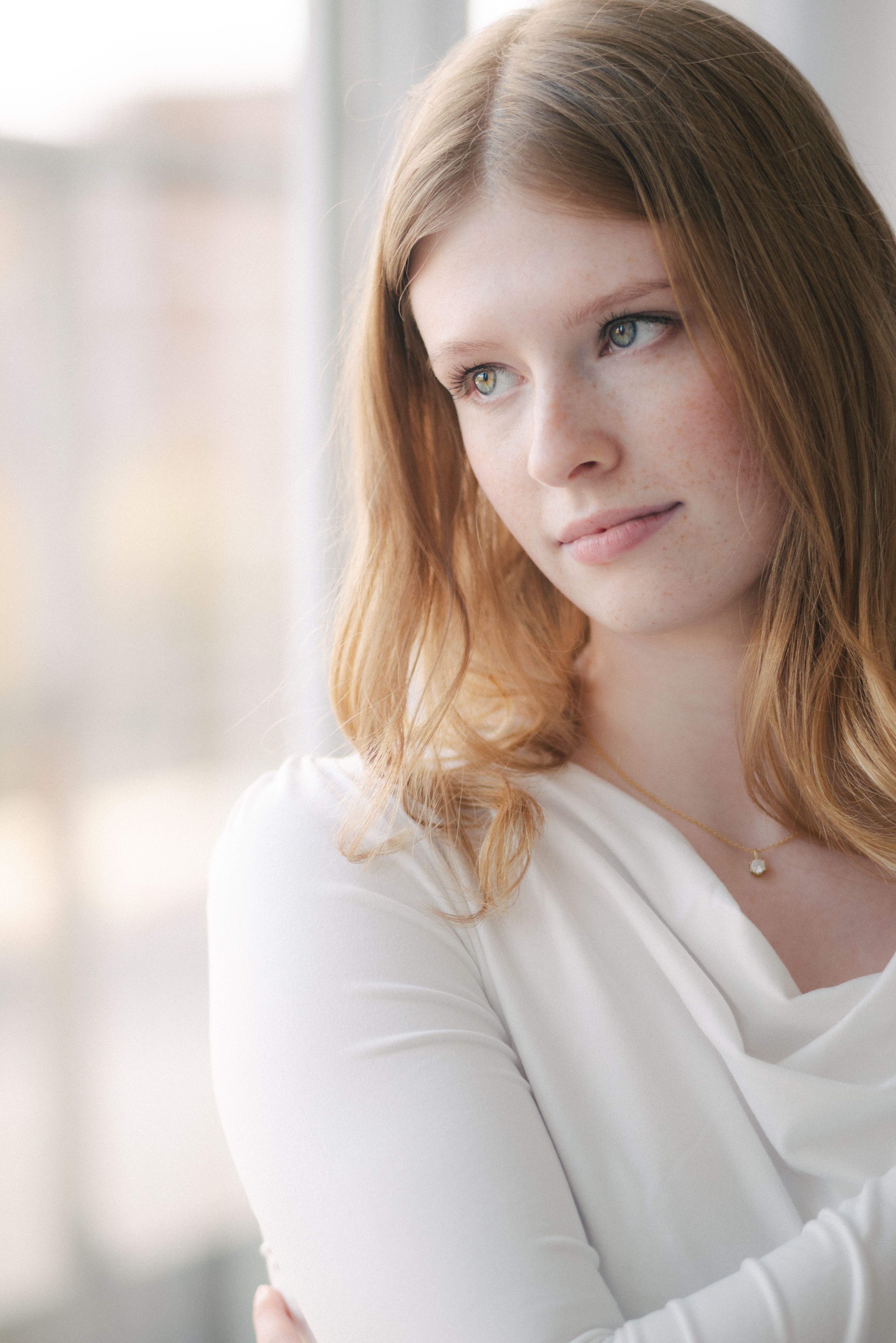 A young woman gazes thoughtfully out of a window, capturing a reflective moment indoors.