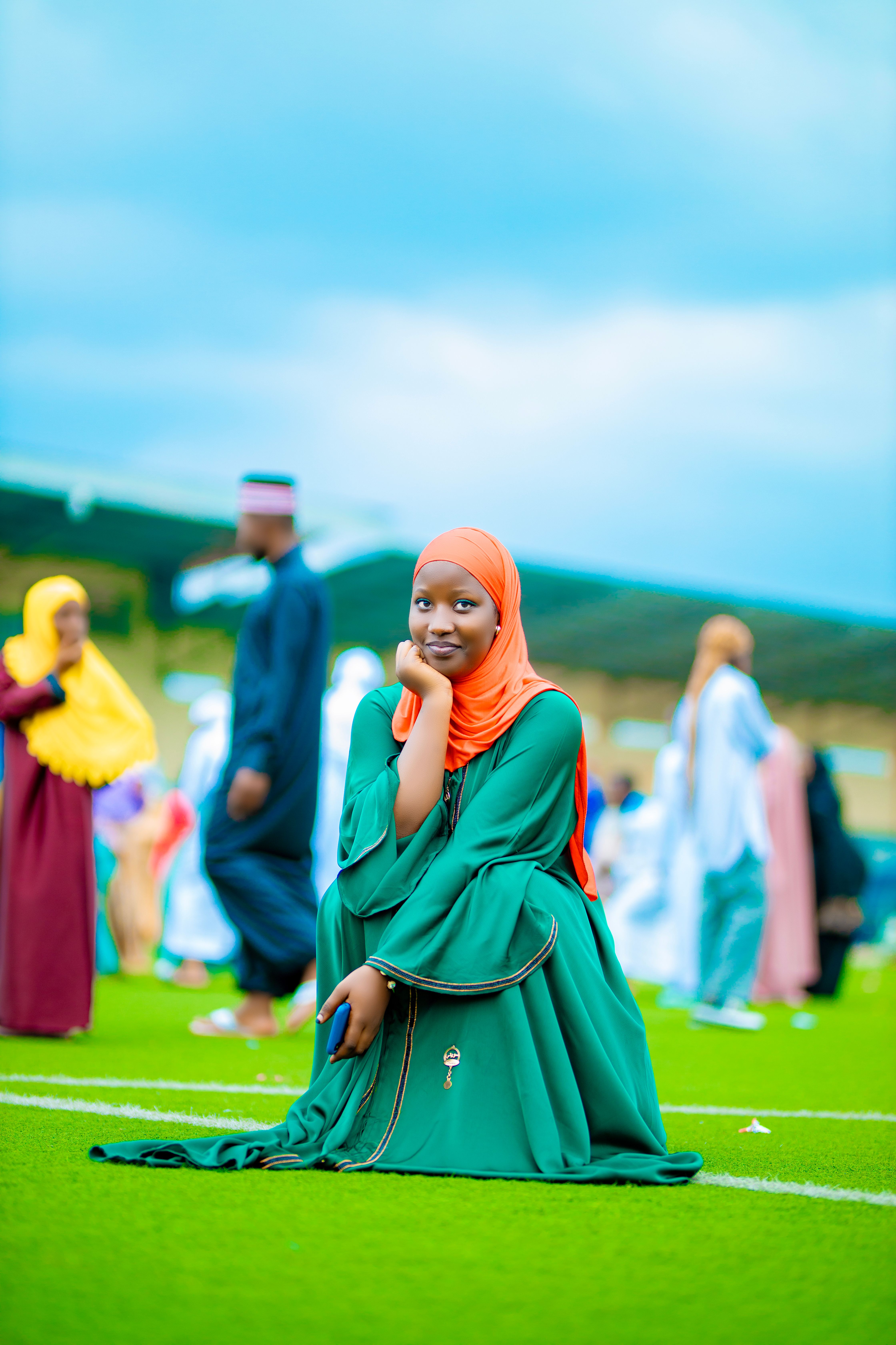A cheerful woman kneeling on grass wearing a colorful hijab in a lively outdoor gathering.