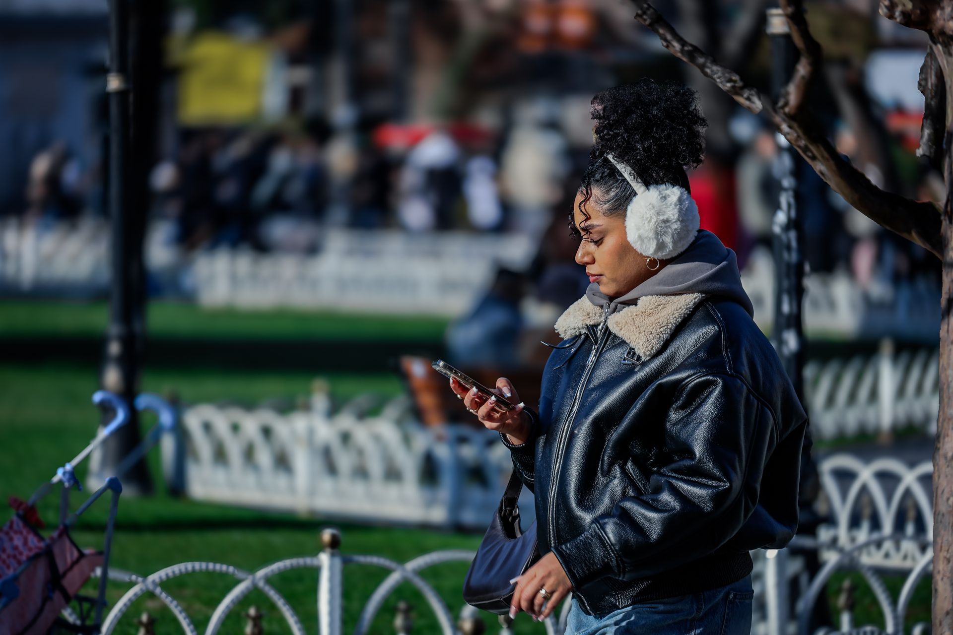 A woman wearing ear muffs and a leather jacket, browsing her phone in an urban park on a sunny day.