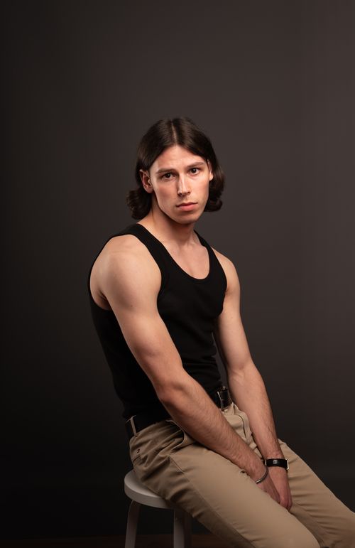 Free Studio portrait of a young man with long hair wearing a black tank top and beige pants, seated on a stool. Stock Photo