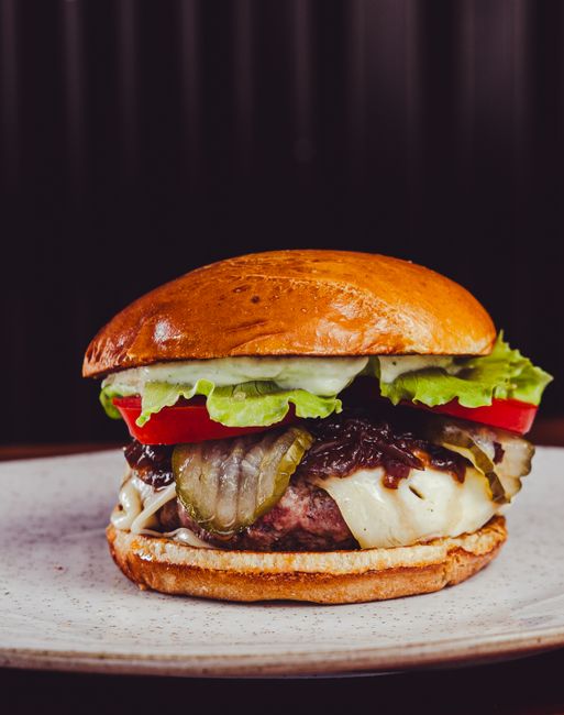 A stacked burger on a sesame seed bun with a charred beef patty, melted cheese, lettuce, tomato slices, pickles and raw onion visible from the side, cut in half on a wood board.