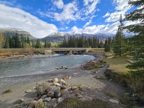 A picturesque autumn view showcasing a serene river, bridge, and snow-capped mountains.