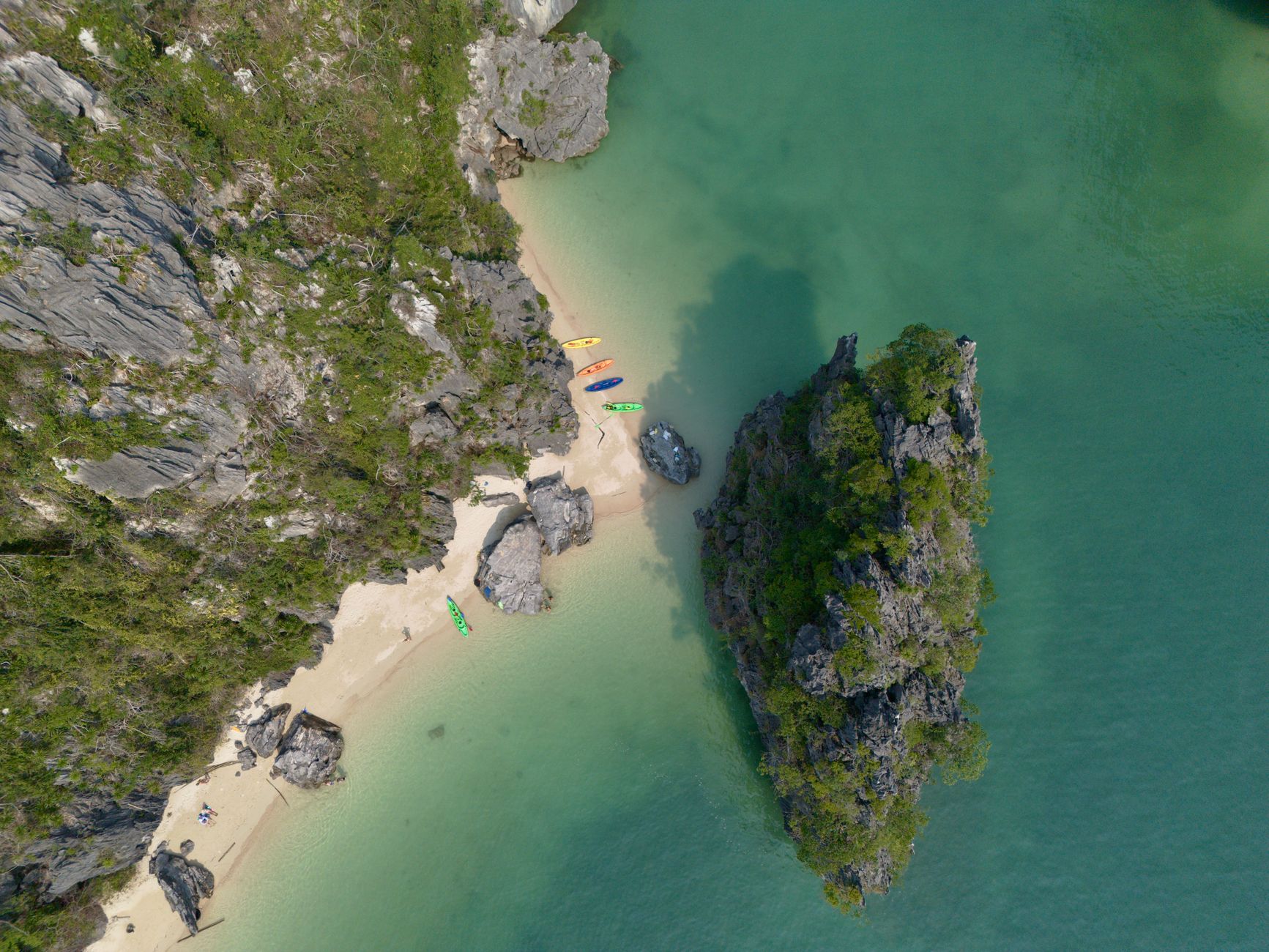 Scenic aerial view of a tropical beach with colorful kayaks, clear waters, and lush green rock formations.