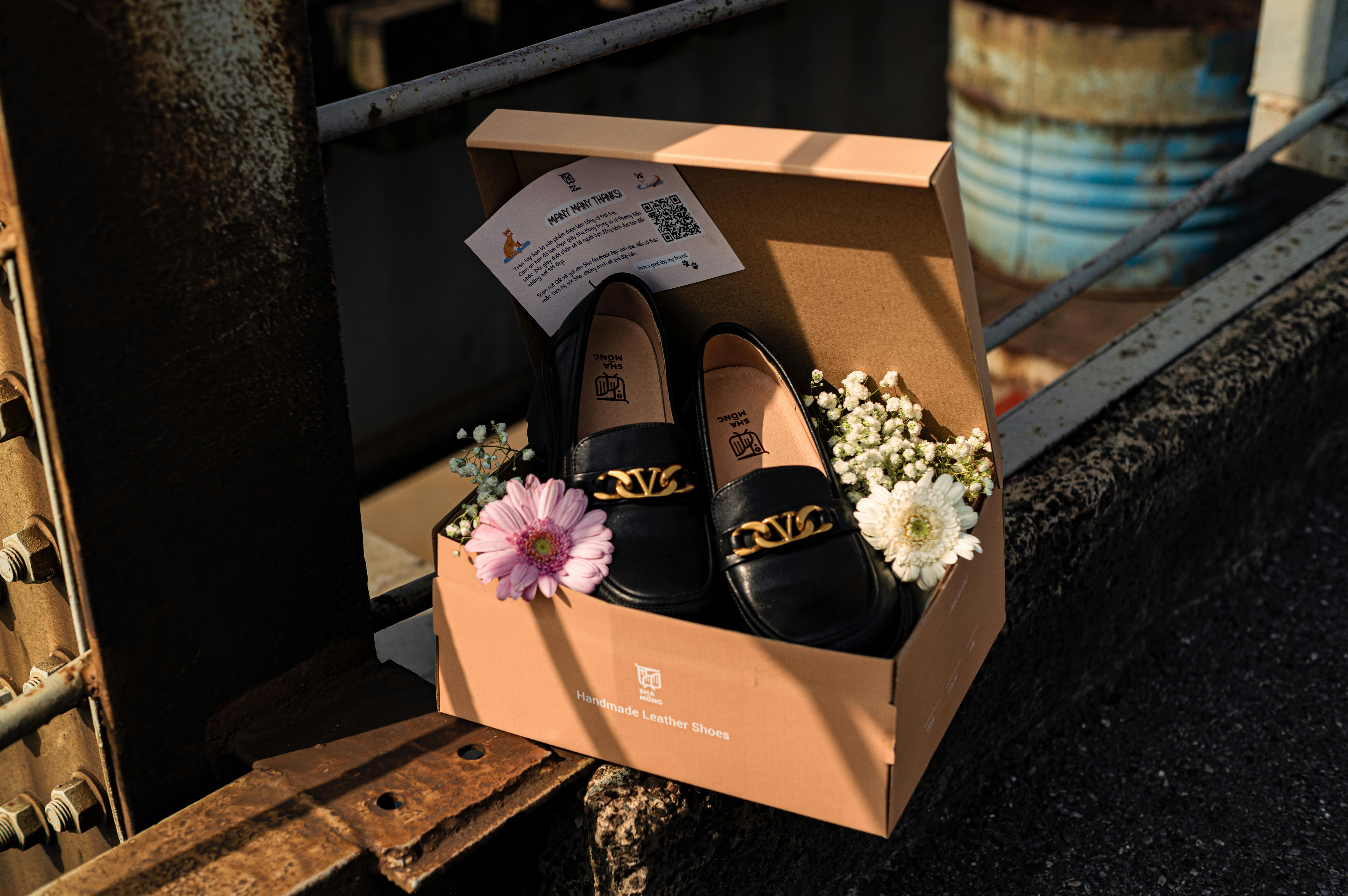 Black leather shoes with gold chain in box, adorned with flowers, in an industrial setting.