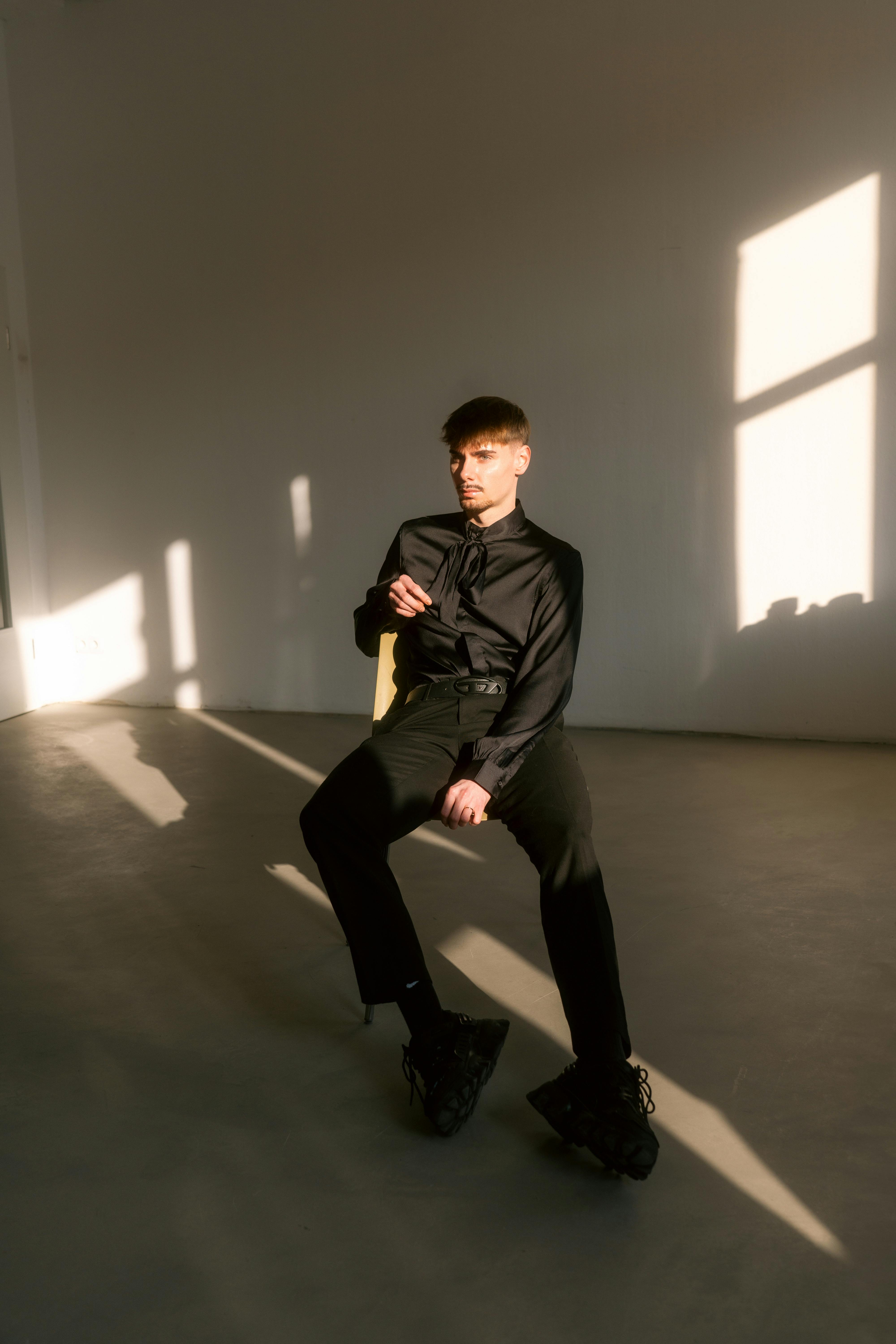 A man in black sits on a chair in a sunlit room, casting long shadows.