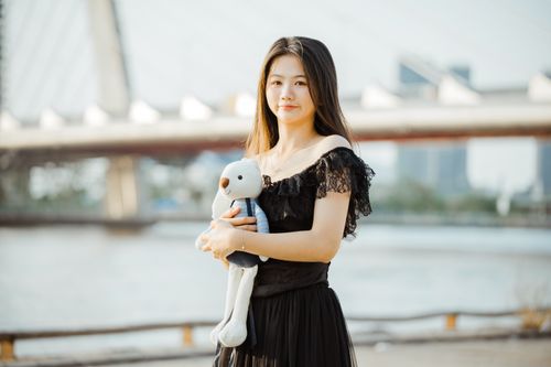 Free Young woman in black dress holding a teddy bear, standing by a bridge on a sunny day. Stock Photo