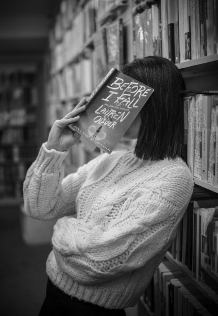 Black and white portrait of a woman hiding behind a book in a library setting.