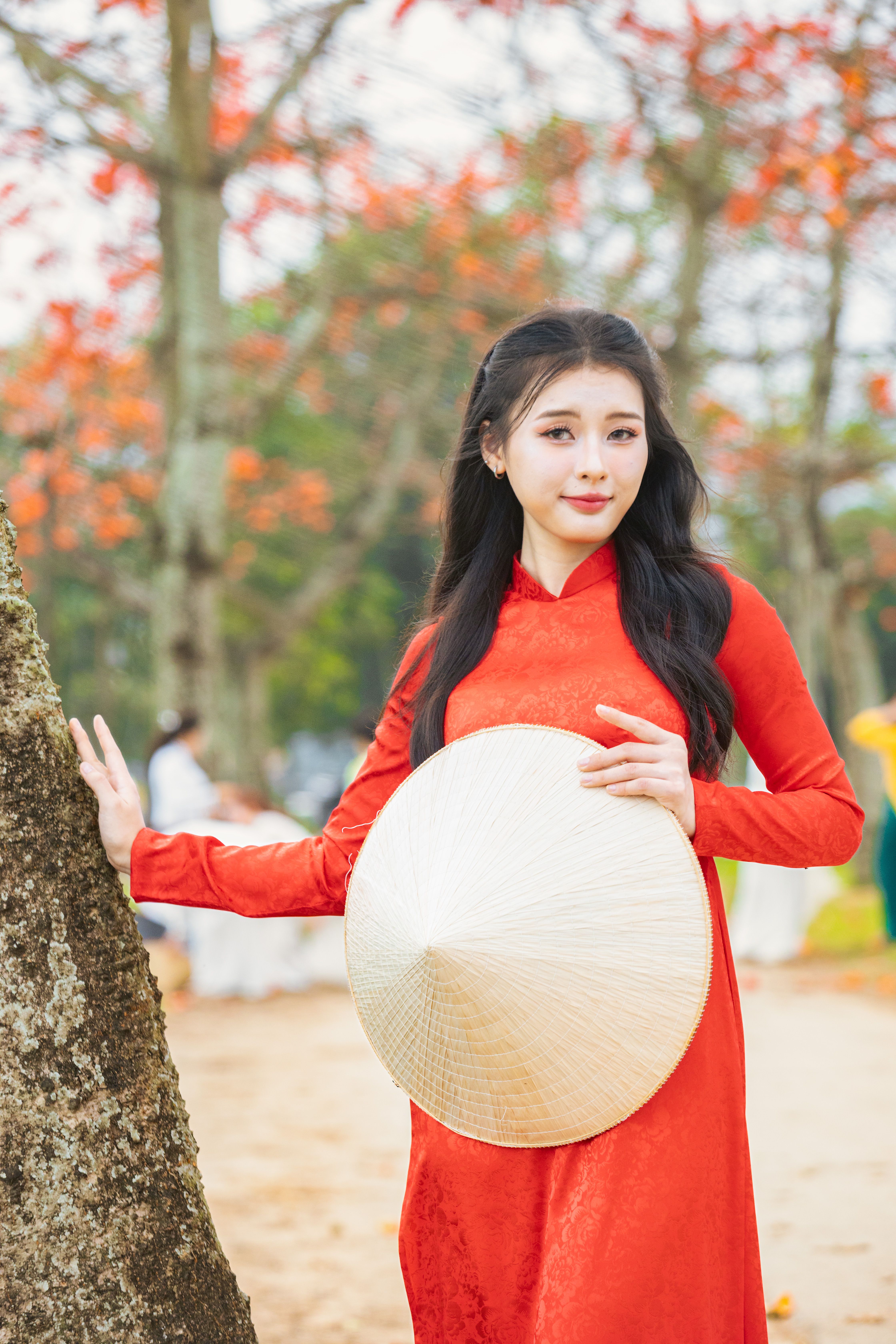 Portrait of a woman in a red Ao Dai holding a conical hat in a serene outdoor setting.