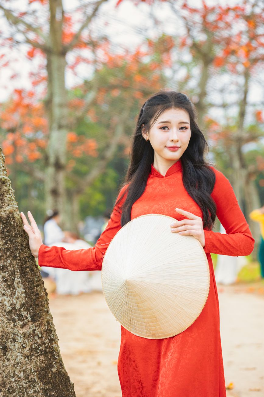 Portrait of a woman in a red Ao Dai holding a conical hat in a serene outdoor setting.