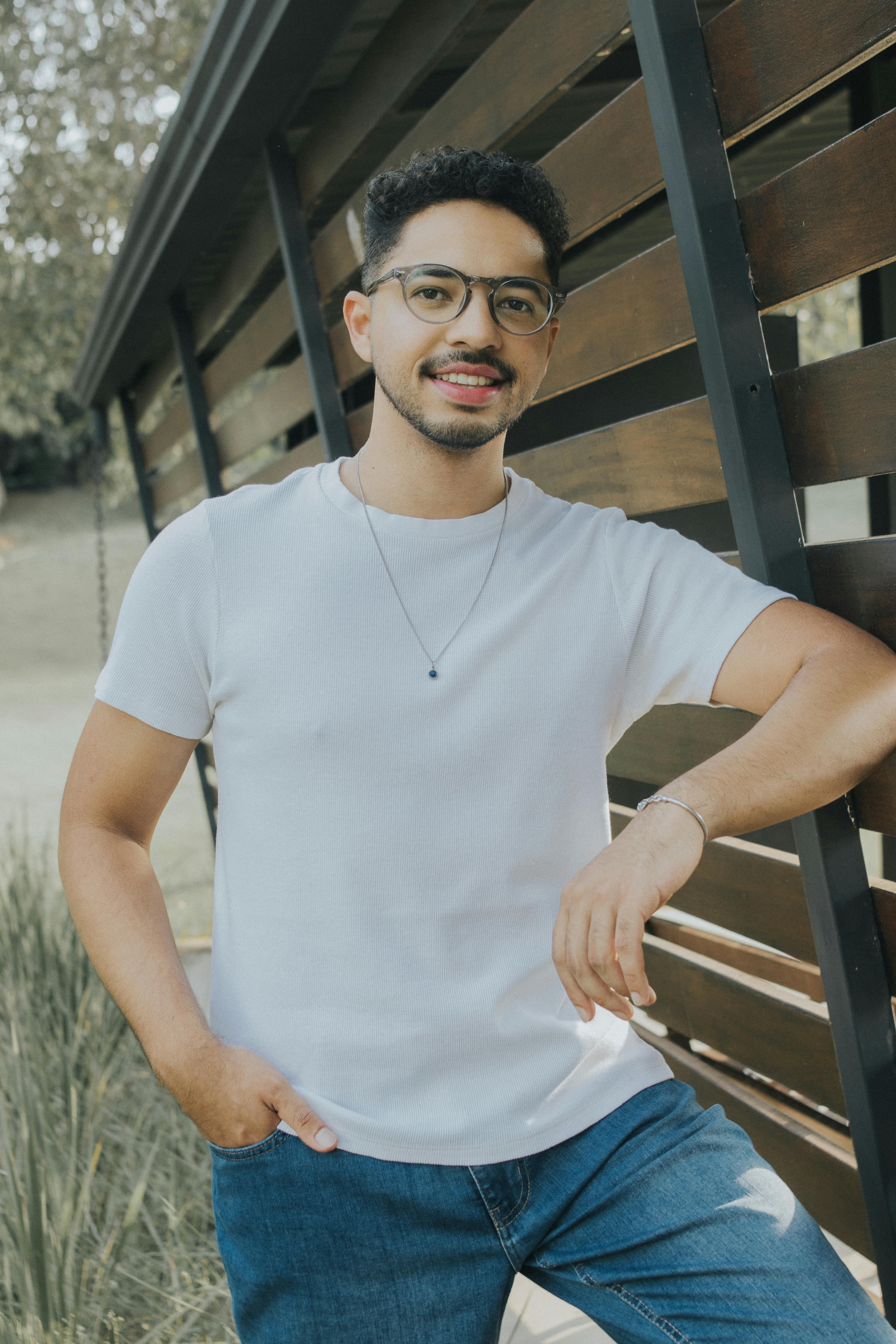 Casual outdoor portrait of a smiling man leaning on a wooden fence, wearing glasses and a white t-shirt.