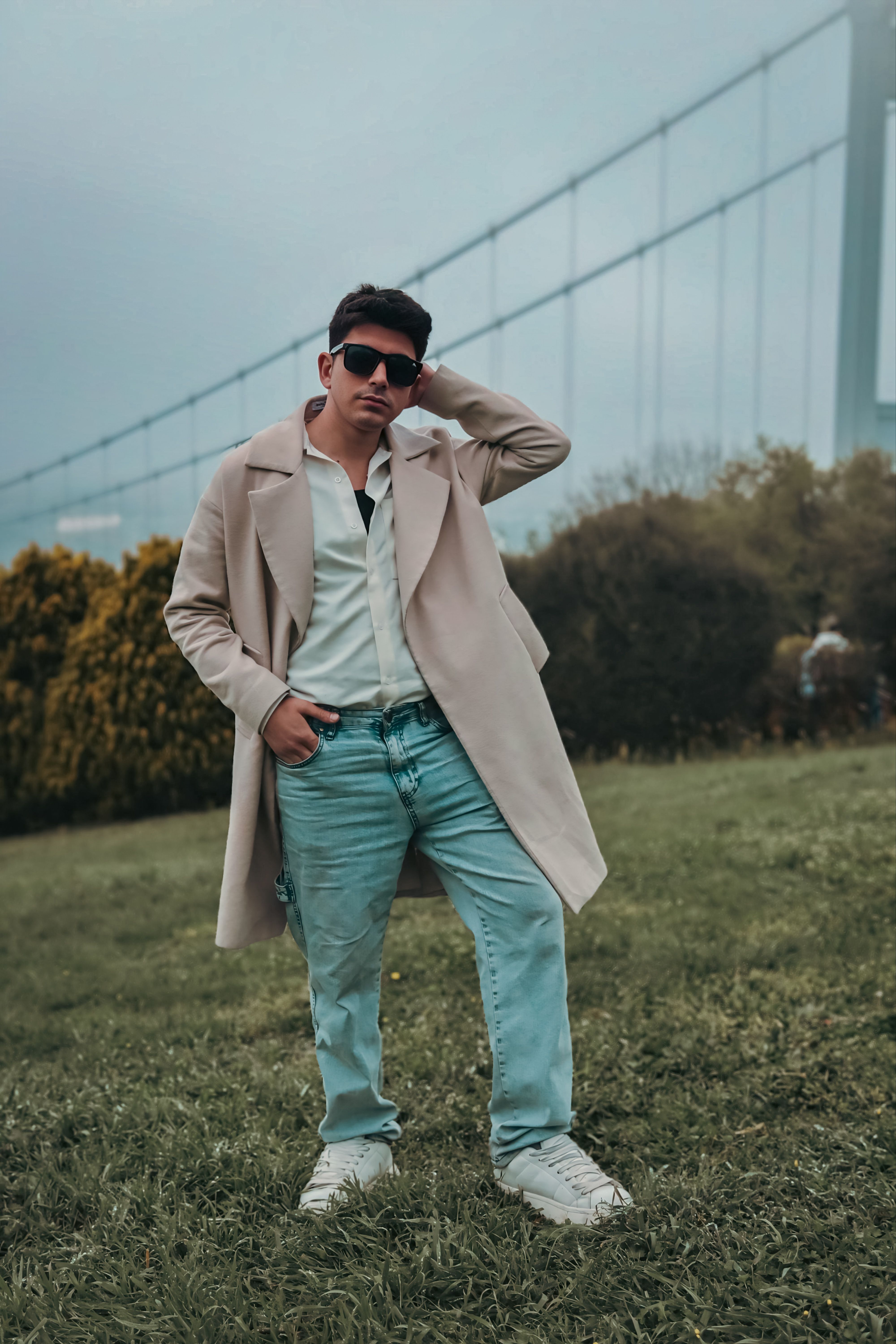 A fashionable young man poses confidently in a scenic outdoor setting near a large bridge.