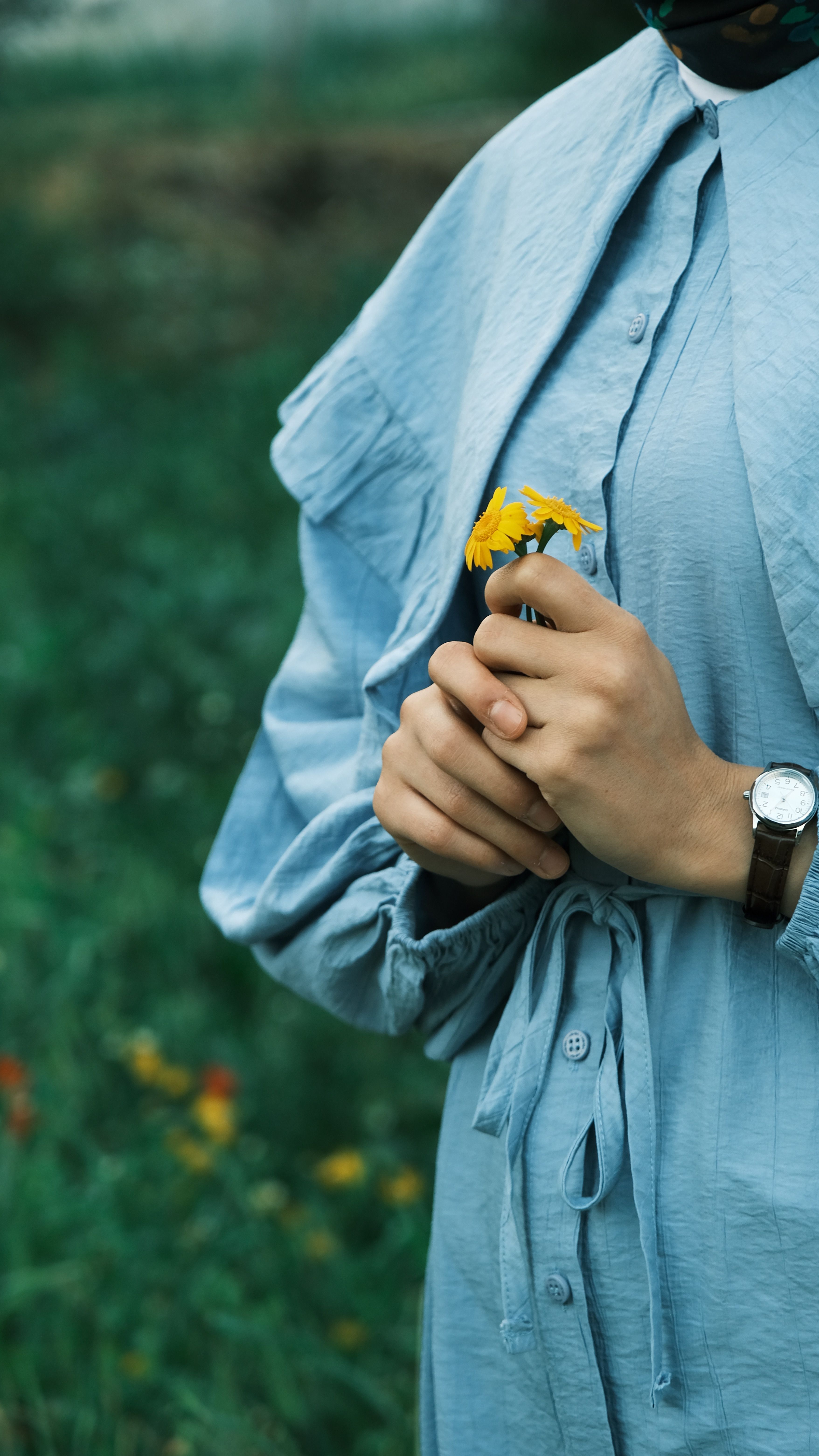A close-up of a person in a blue dress holding a yellow flower outdoors, evoking a serene mood.