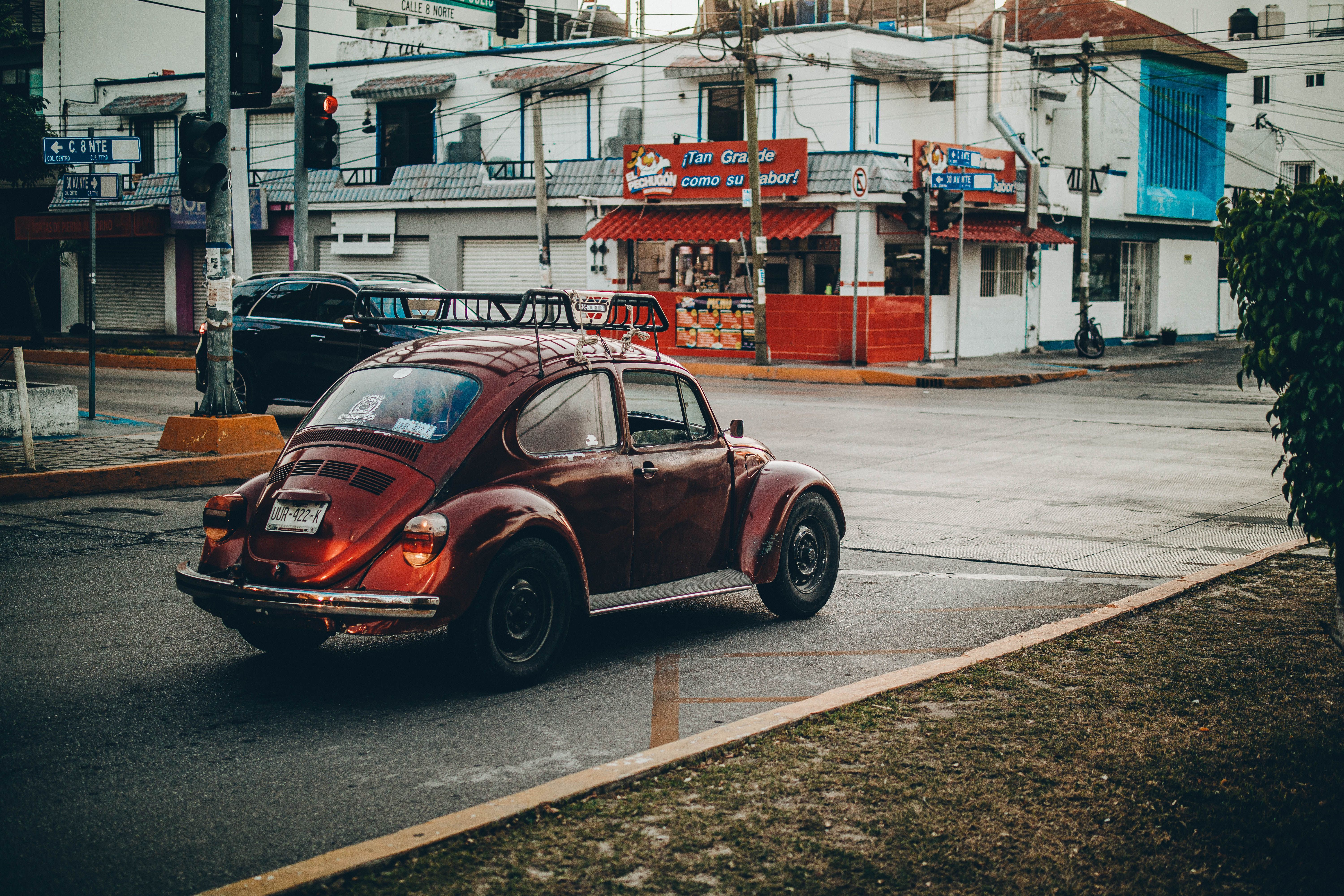 A vintage red Volkswagen Beetle driving through a quiet urban street corner during daytime.