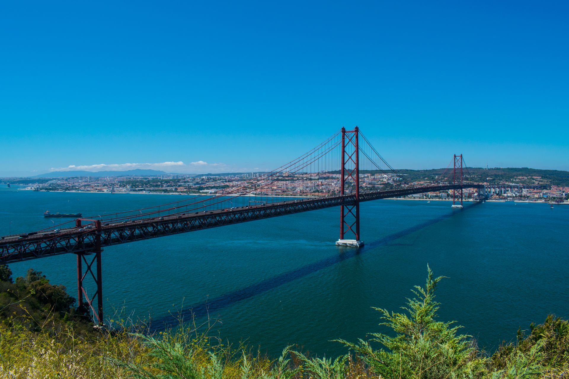 Aerial view of the iconic 25 de Abril Bridge across Lisbon's Tagus River on a clear summer day.