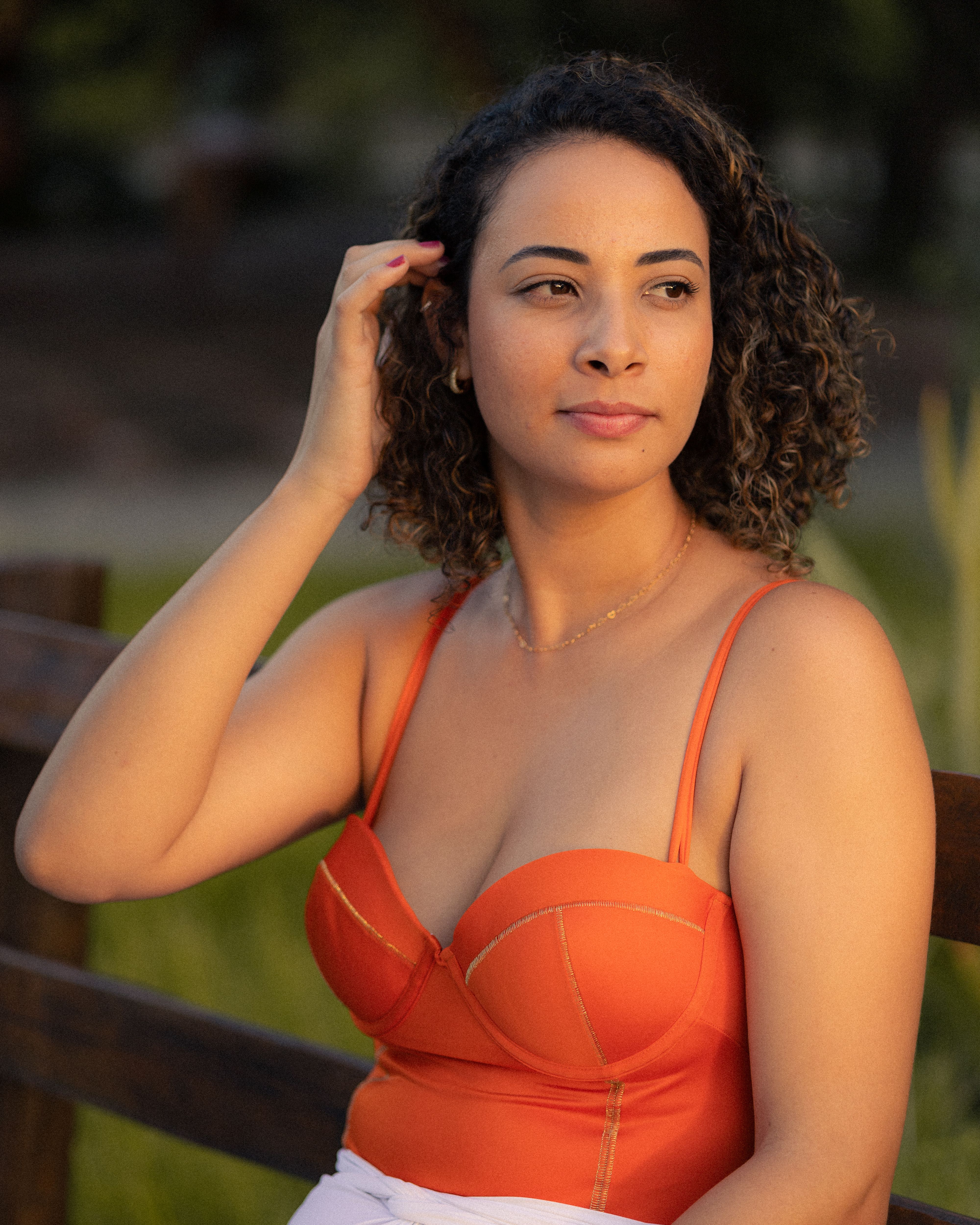 A woman in an orange top poses thoughtfully outdoors during golden hour, capturing warm and natural light.