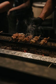 Chef grills assorted skewered meats over charcoal in a dimly lit restaurant setting.