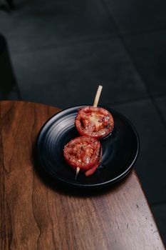 Close-up of grilled tomato skewers on a black plate placed on a wooden table.