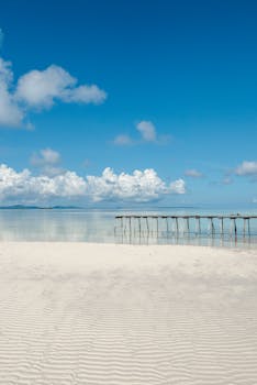 A serene coastal landscape featuring a clear blue sky, azure sea, and sandy beach with a wooden pier.