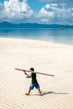 A child walks across a serene beach with clear blue waters and mountains in the background.